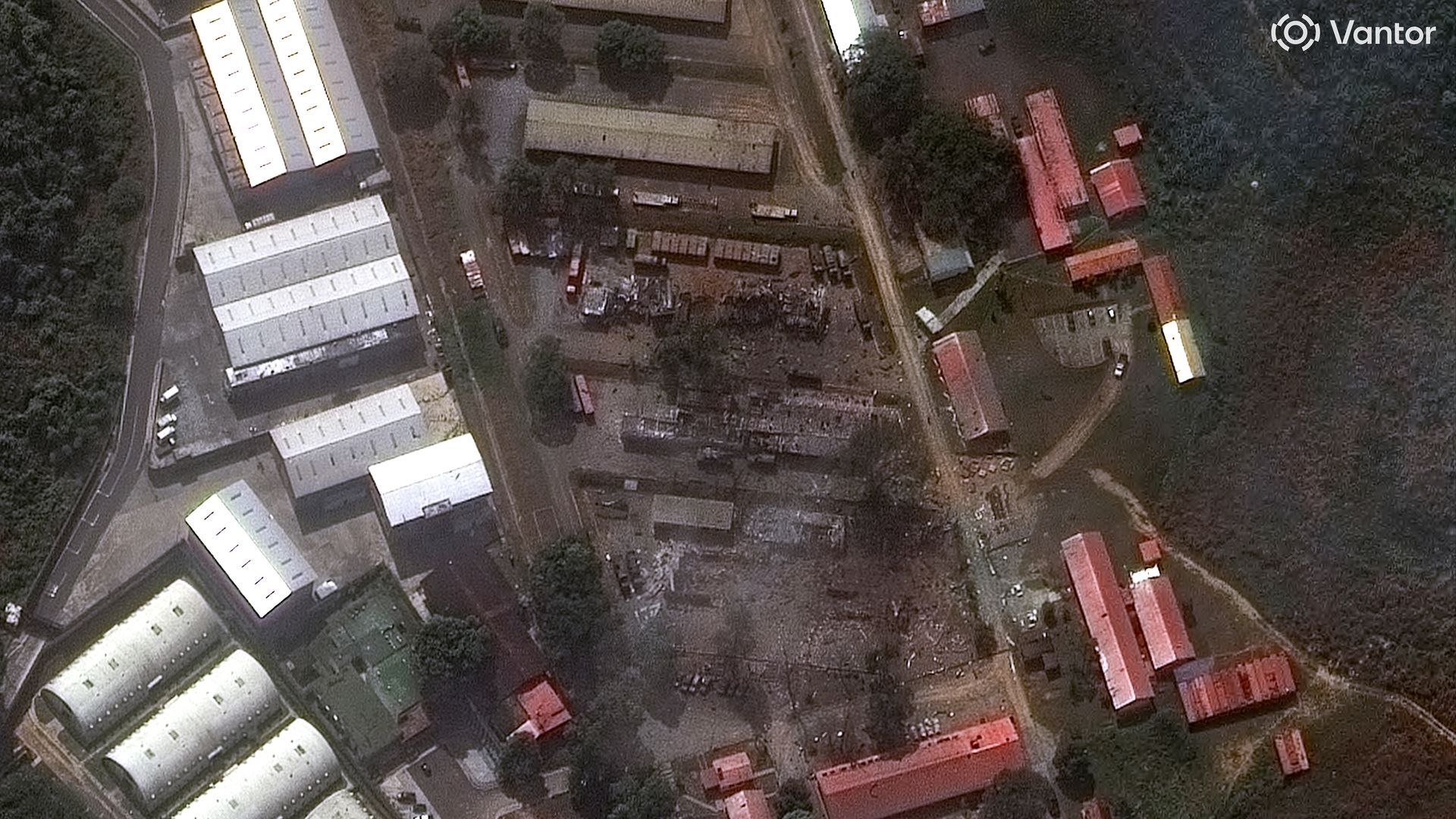 Satellite view of an industrial site with multiple white and red-roofed buildings, roads, trees, and an open area with scattered debris or equipment in the center.
