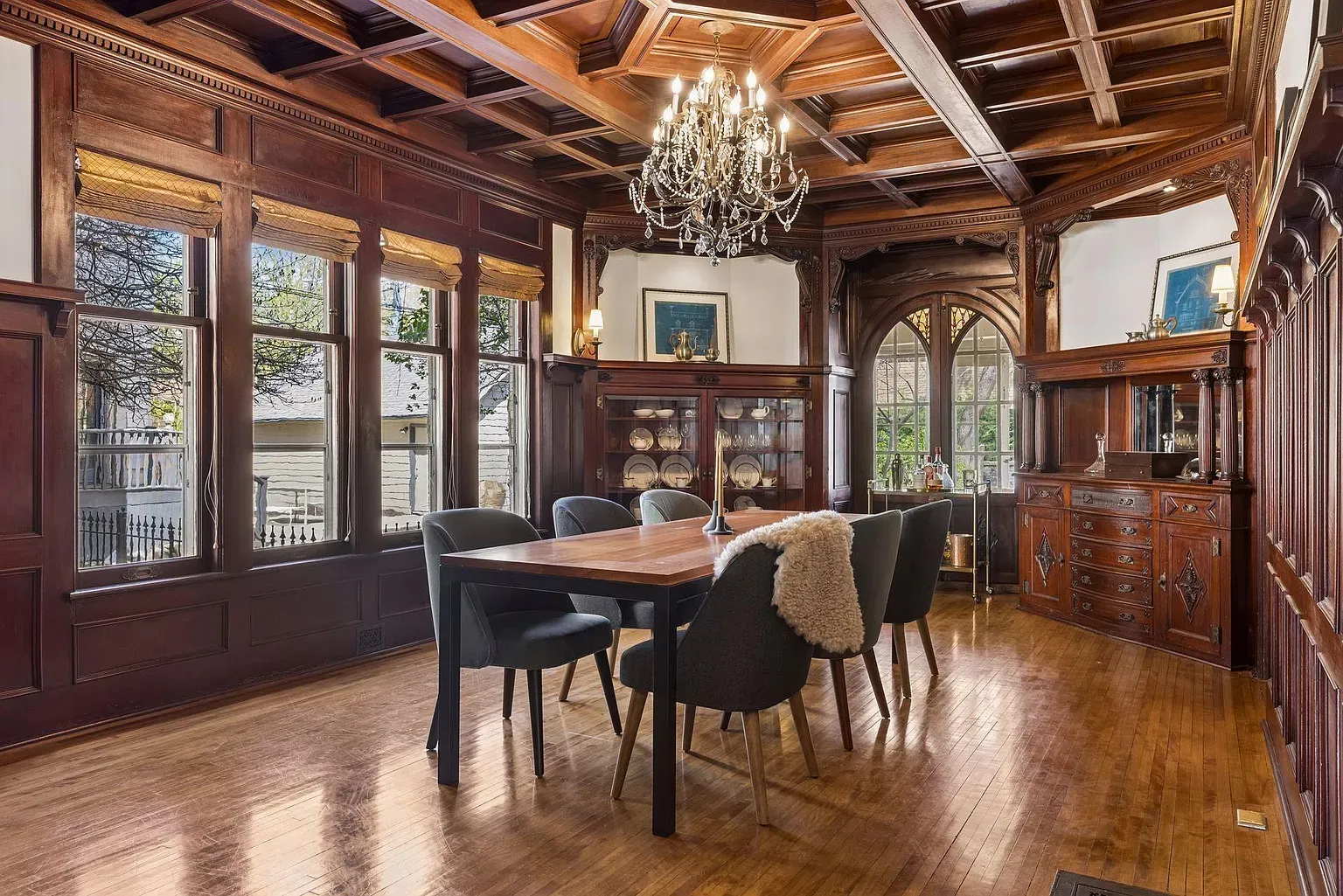Dark wood dining room with a coffered ceiling and crystal chandelier. A wooden table with six upholstered chairs, a fur throw, and a china cabinet by paneled walls and tall windows.