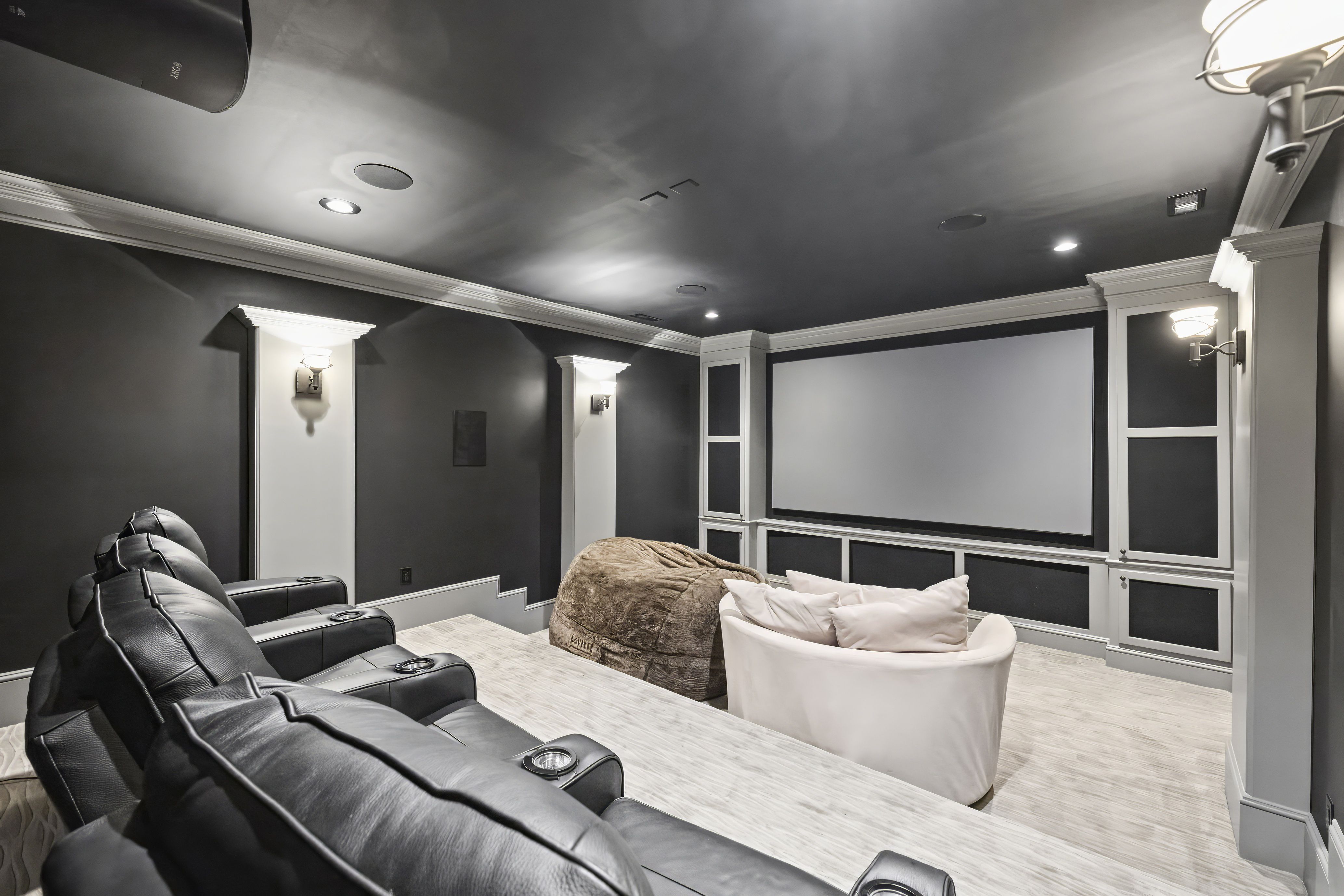 Dark gray home theater with white trim and built-in cabinets. Black leather recliners line the left, white chairs in front, and a large projection screen on the back wall; ceiling lights.