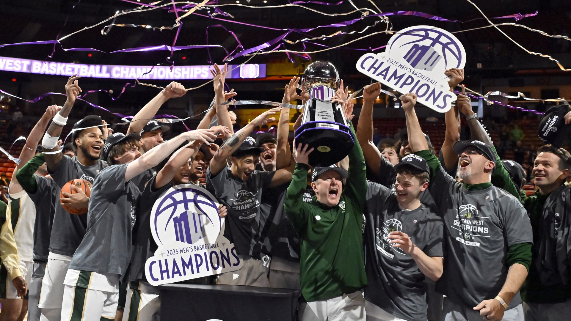 A man in a green jacket lifts a giant trophy surrounded by players holding up signs that say CHAMPIONS.