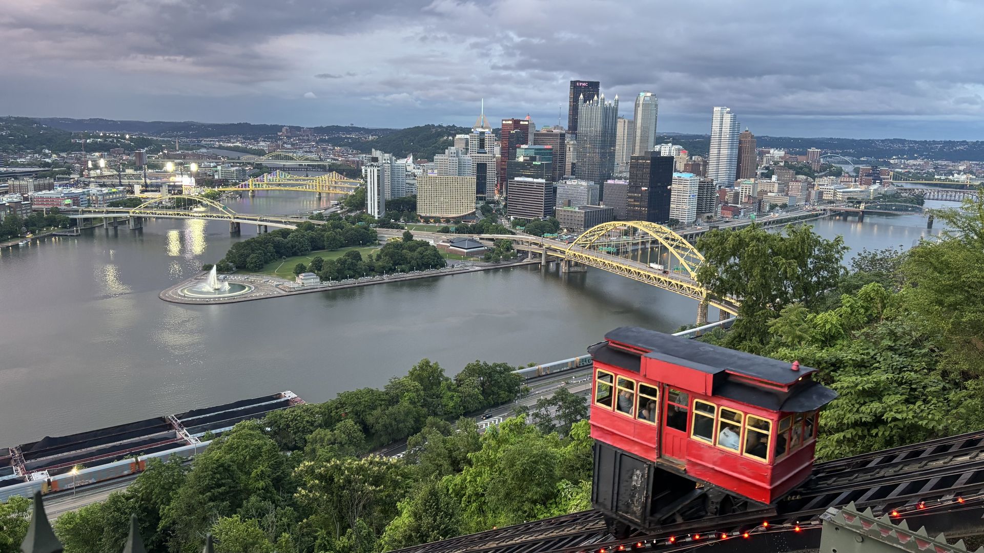 View of Pittsburgh skyline with tall glass towers, yellow bridges across the rivers, a circular fountain, green riverfront park, and a red incline trolley car in the foreground on the hill.