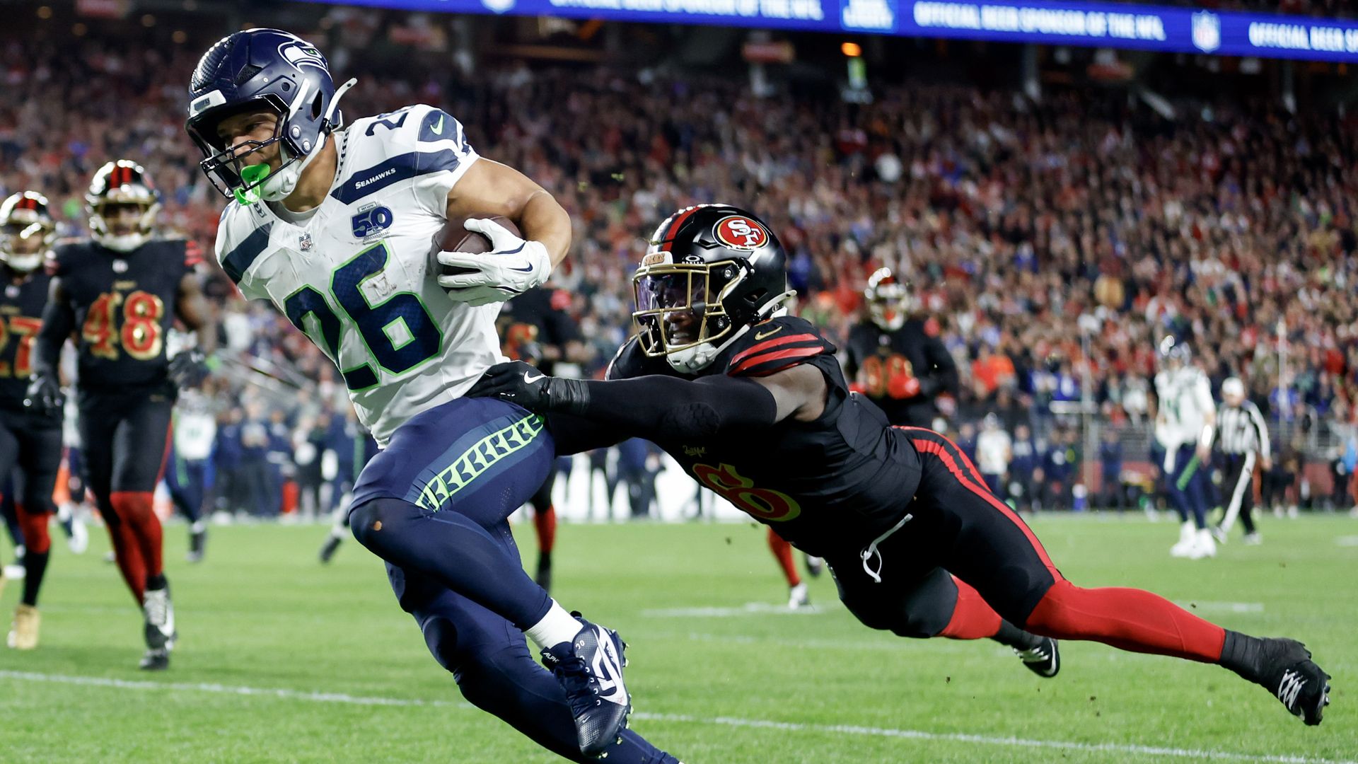 Seattle Seahawks player #26 runs with football as San Francisco 49ers player #30 dives trying to tackle him during a night game with a crowded stadium.