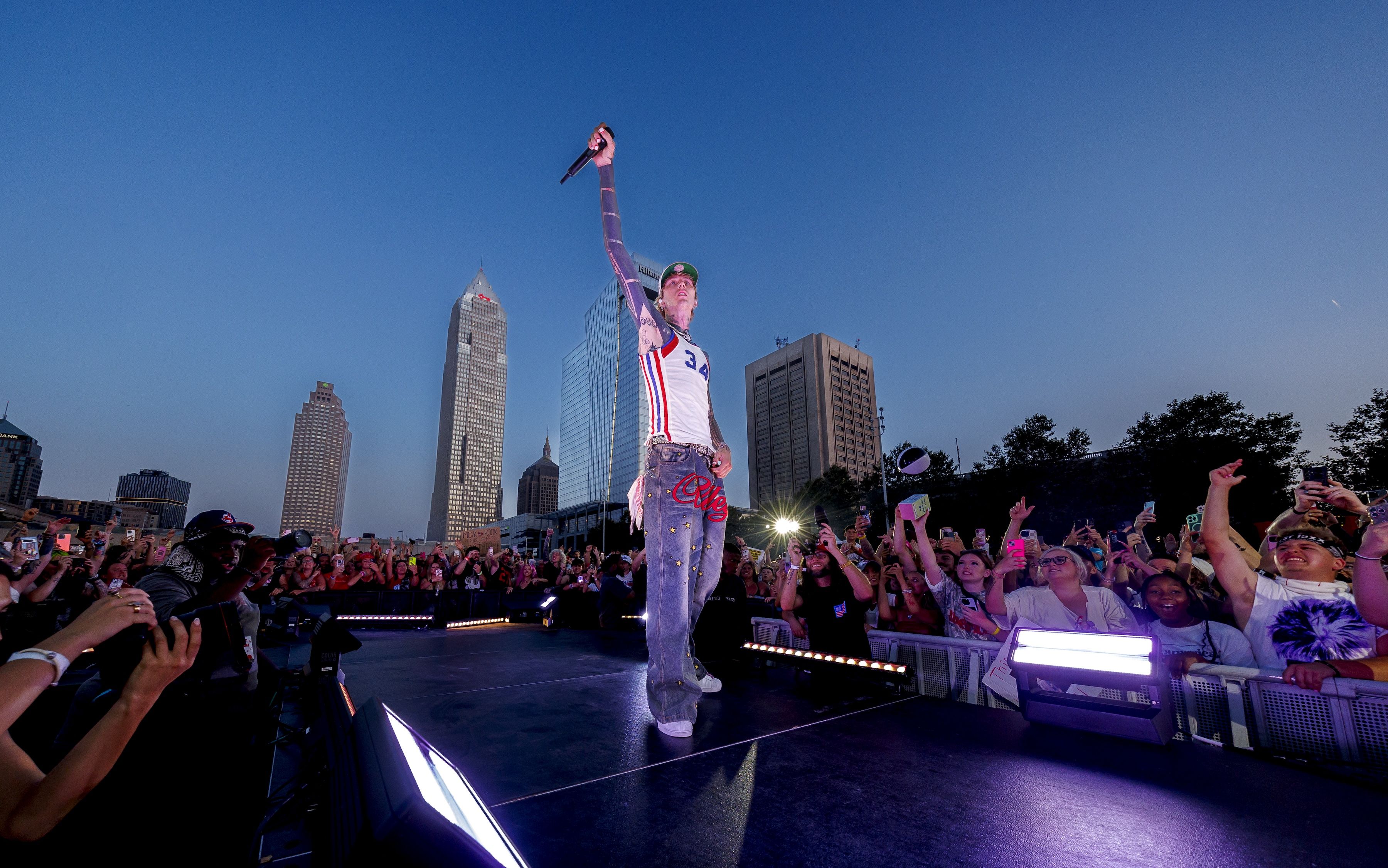 Machine Gun Kelly in jeans and a white tank top with number 34 performs on outdoor stage at dusk, holding a microphone up, with a large, enthusiastic crowd and city skyscrapers in the background.