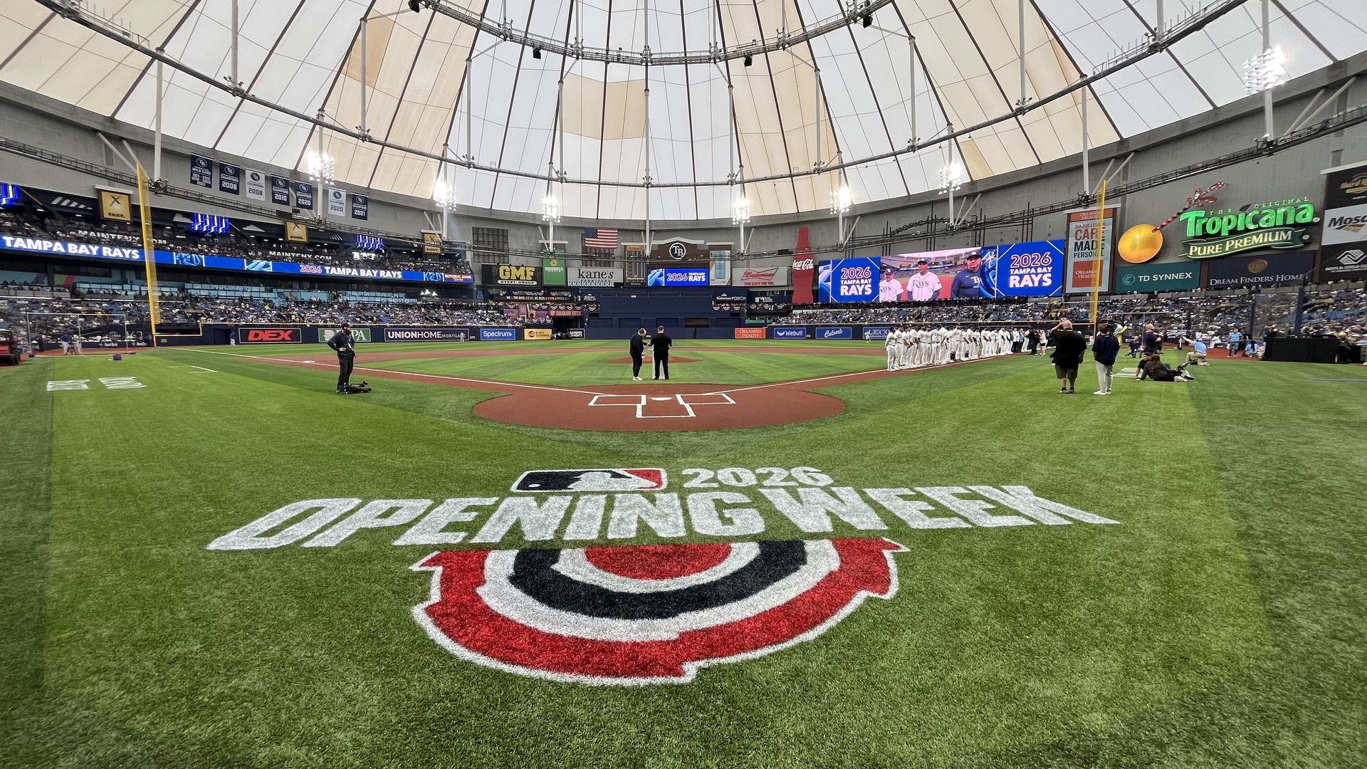 Indoor baseball field inside a domed stadium; a large "2023 Opening Week" logo on the turf. Rays players in white line up on the right as officials near home plate, and banners around.