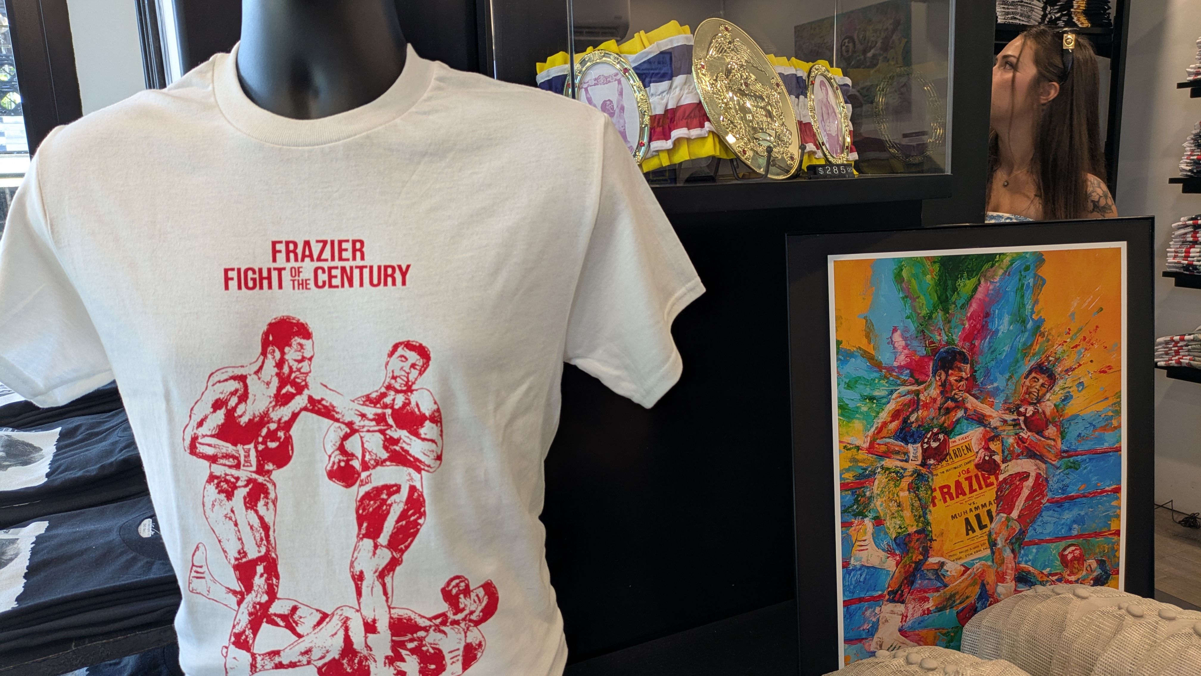 Mannequin wearing a white T-shirt with a red boxing graphic and the words "FRAZIER FIGHT OF THE CENTURY"; background shows a display case with boxing belts and a colorful framed boxing poster, with a woman on the right.