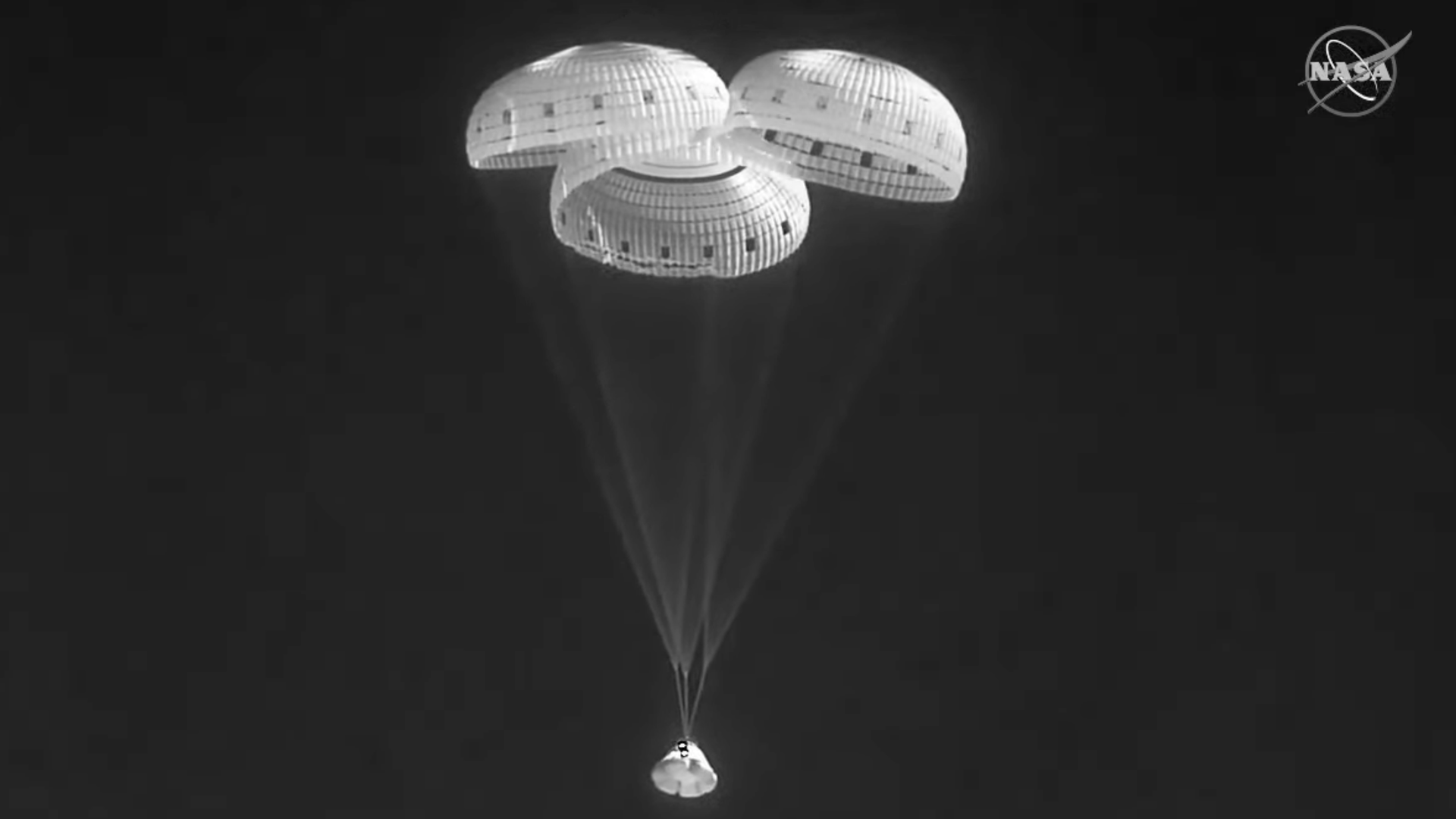 A black and white image of the Starliner coming down to Earth under three parachutes