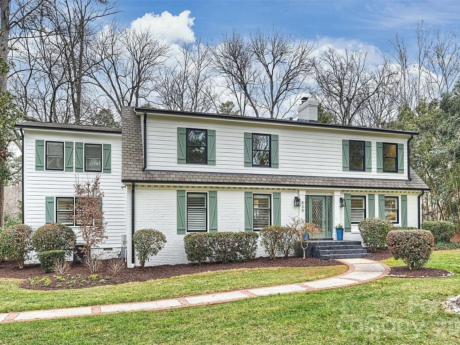 Two-story white house with green shutters and front door, a curved concrete walkway, manicured bushes, and leafless trees in the background under a partly cloudy sky.