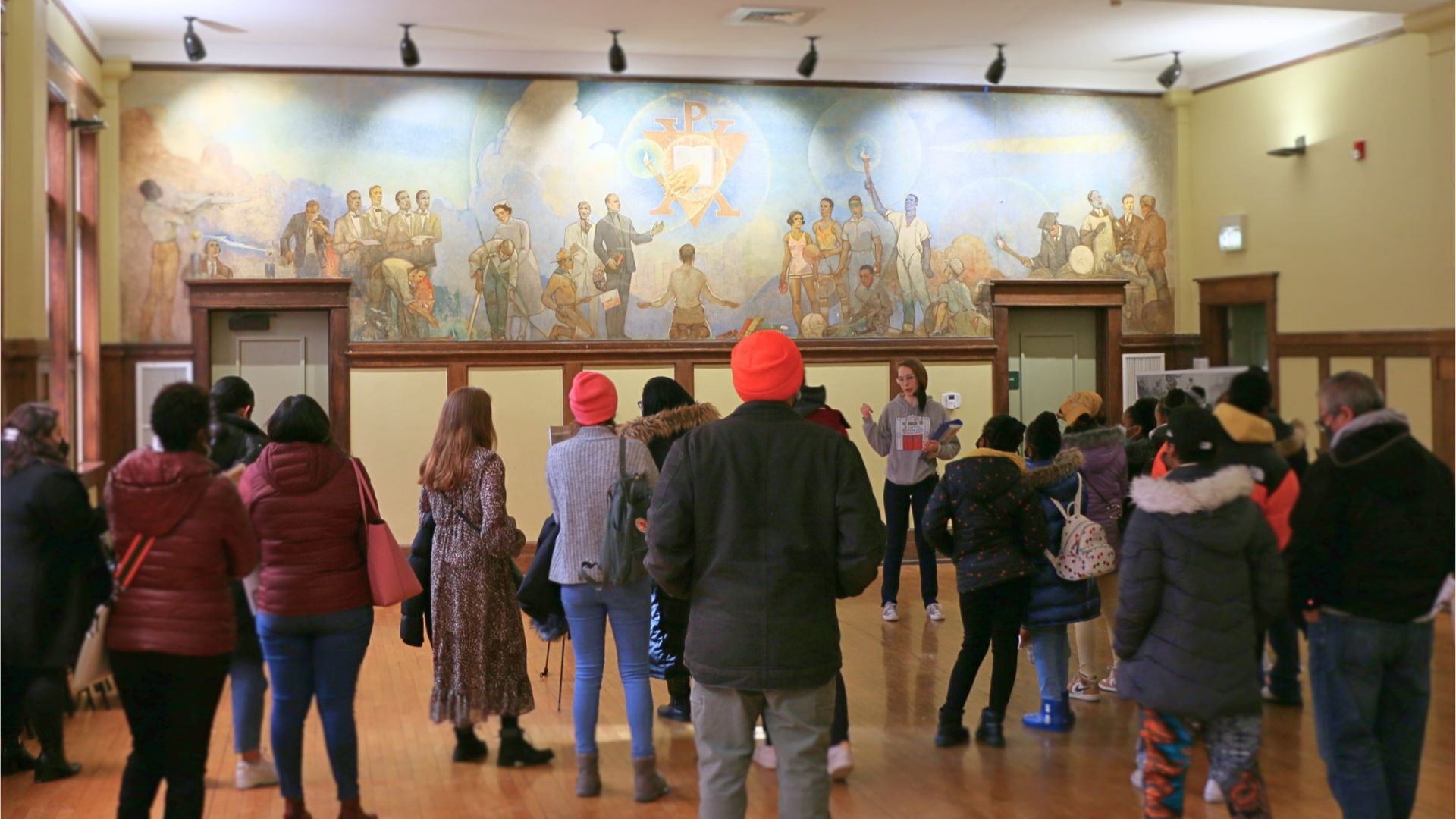 The backs of people listening to a tour guide who's standing in front of a mural.