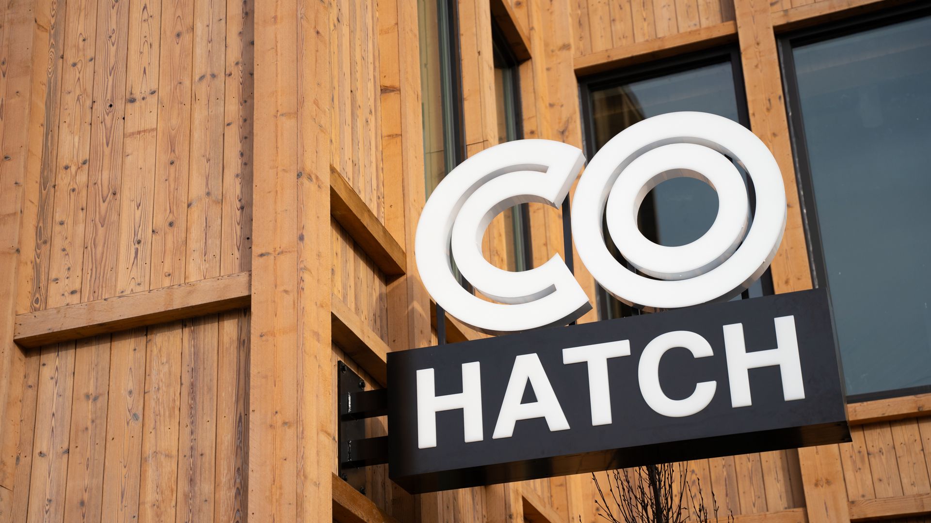 White and black sign reading "CO HATCH" mounted on a natural wood-paneled building exterior with large windows.