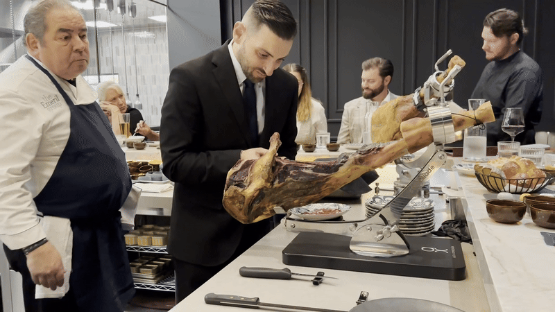 Emeril Lagasse watches as a man carves a slice of ham off a jamon.