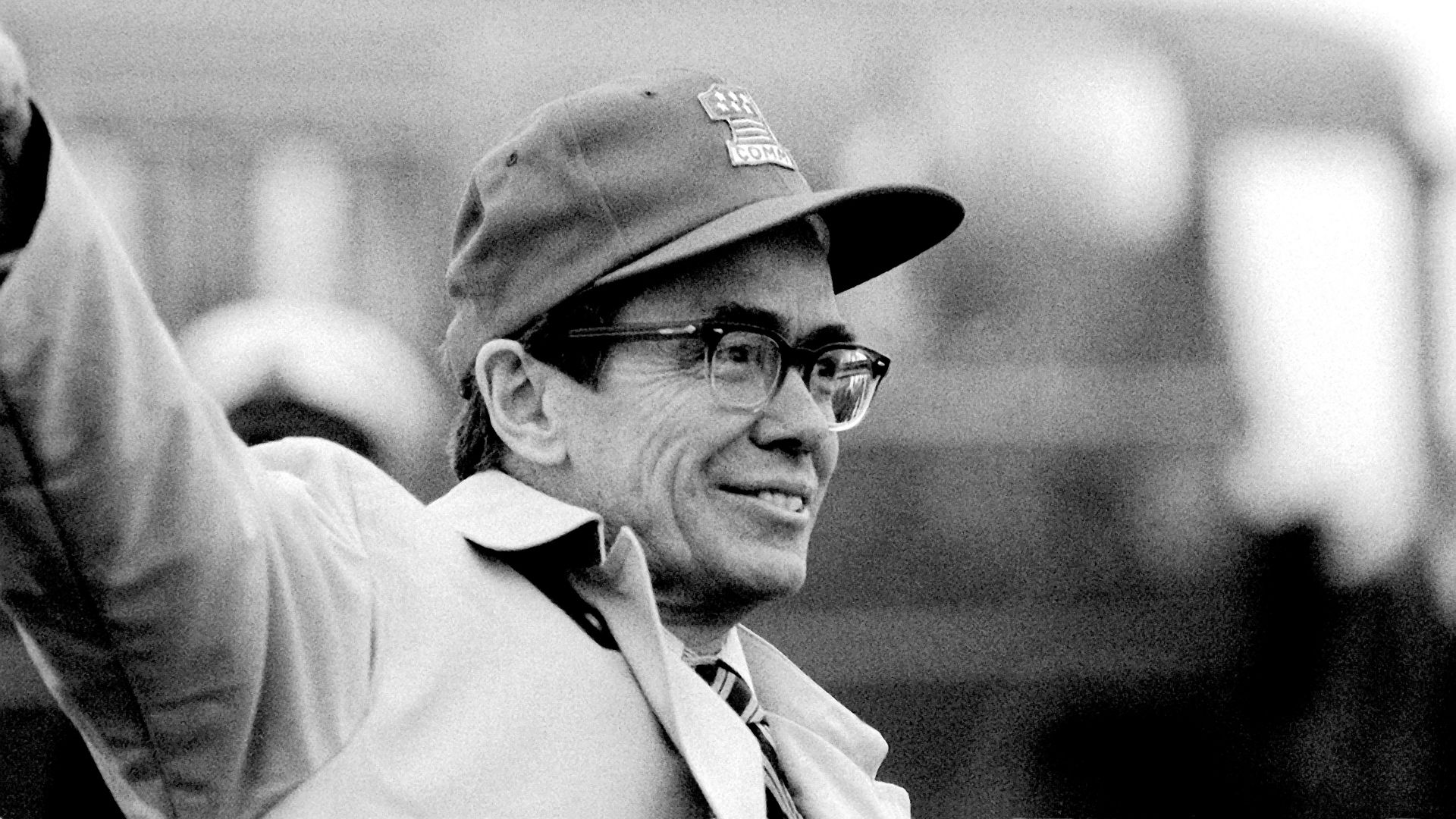 A black and white photo of Bruce Laingen wearing a baseball hat and glasses, waving 