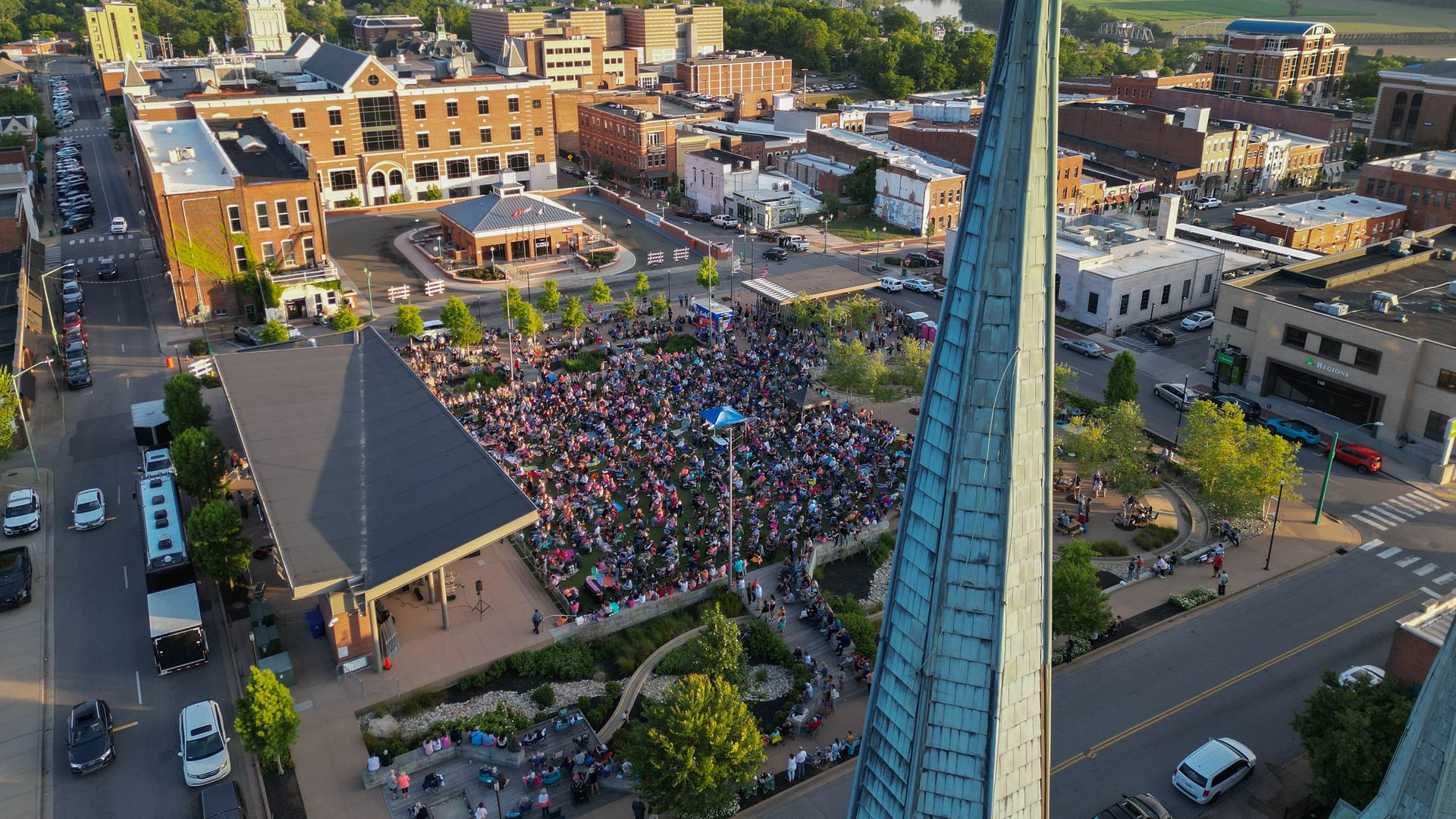 Aerial view of a downtown plaza crowded with people, surrounded by brick buildings, with a tall blue-green spire in the foreground and a river and green fields in the distance.