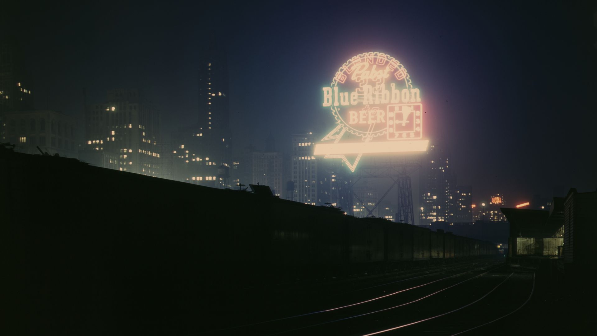 Night cityscape with a dark foreground and distant lit buildings; a bright neon "Blue Ribbon Beer" sign in a circular logo glows, casting purple light over railroad tracks.