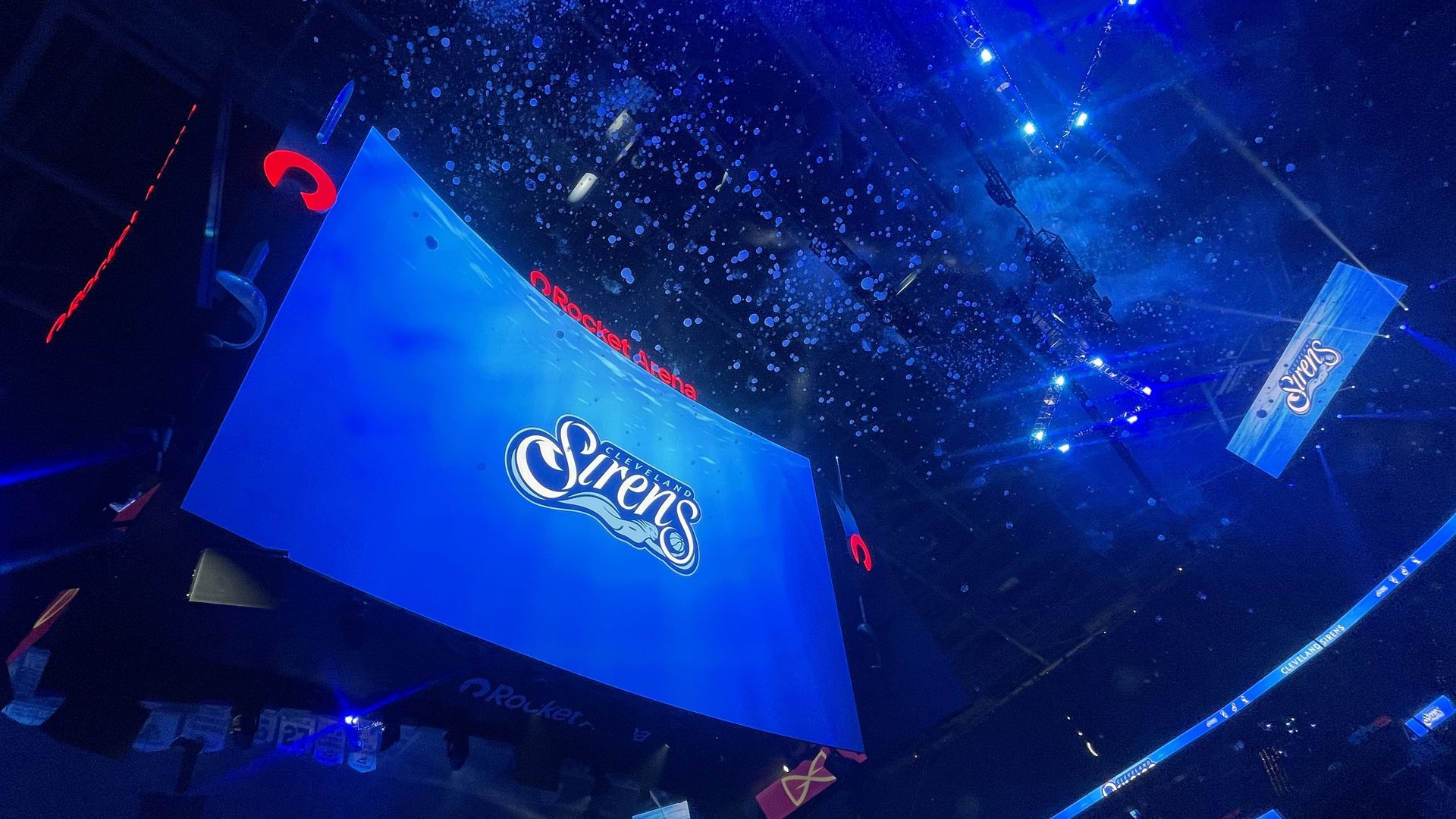 Blue-lit indoor arena with a large blue stage screen, overhead white confetti, and hanging banners along the sides.