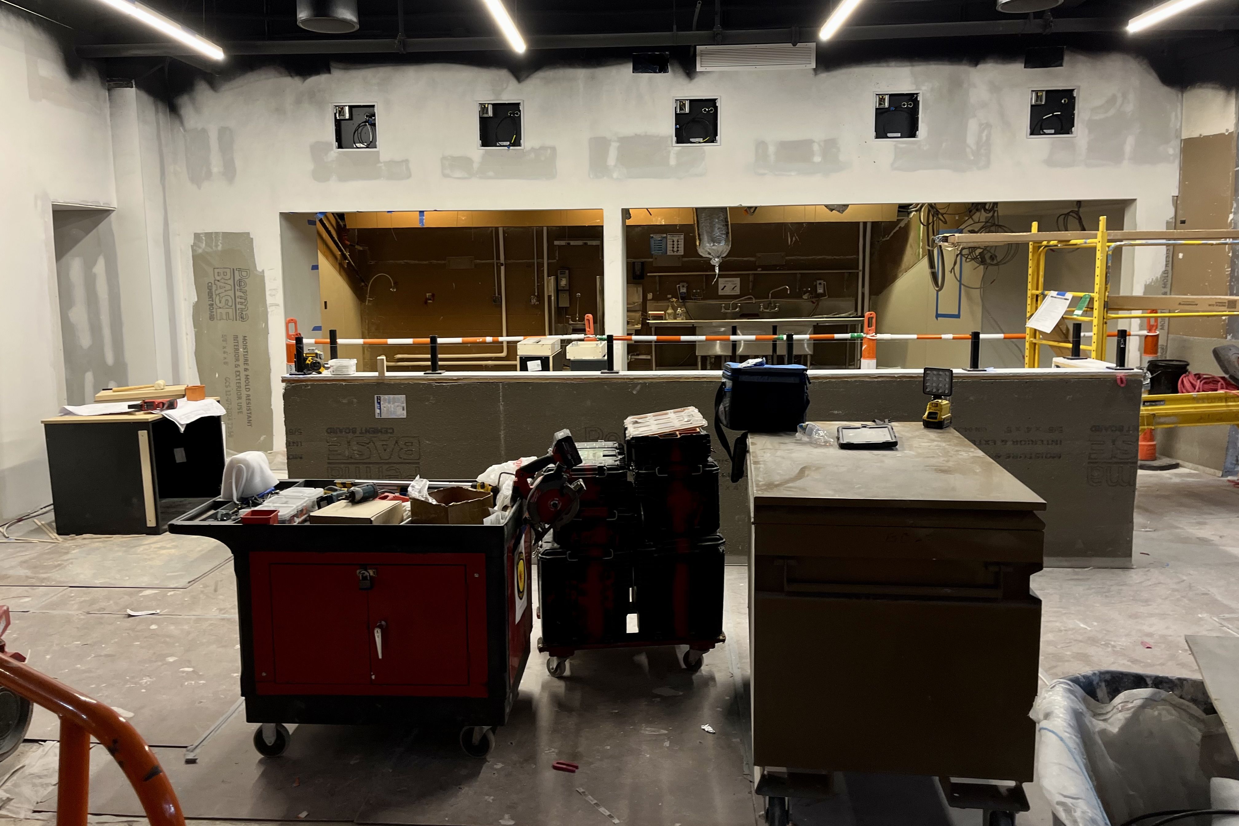 Indoor construction site with ongoing wall work, tools and equipment on carts, orange safety railing, and exposed wiring and pipes visible through unfinished wall openings.