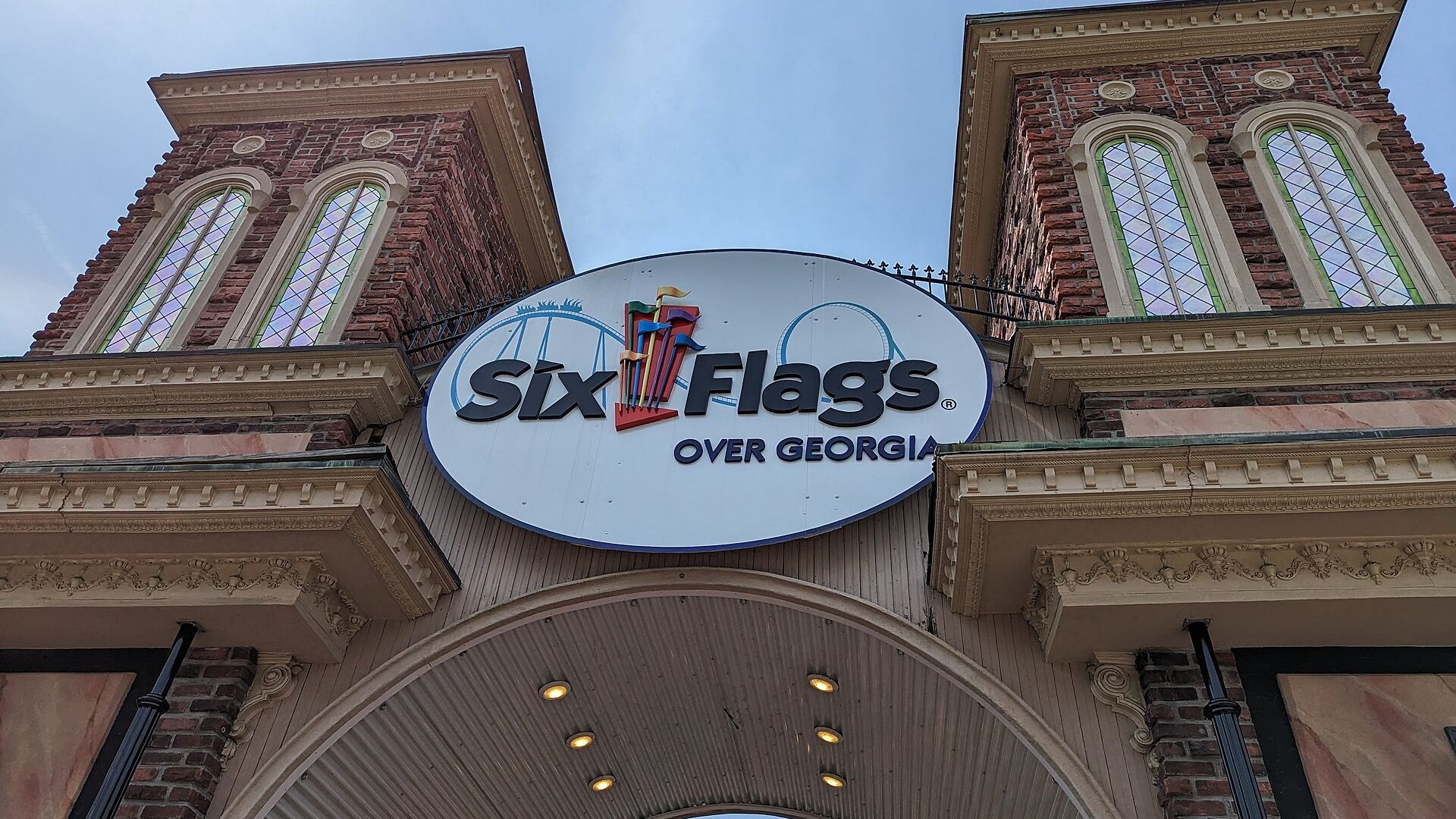 Entrance to Six Flags Over Georgia showing two brick towers with arched green-tinted windows, a white curved canopy, and a large oval sign reading "Six Flags Over Georgia" against a clear blue sky.
