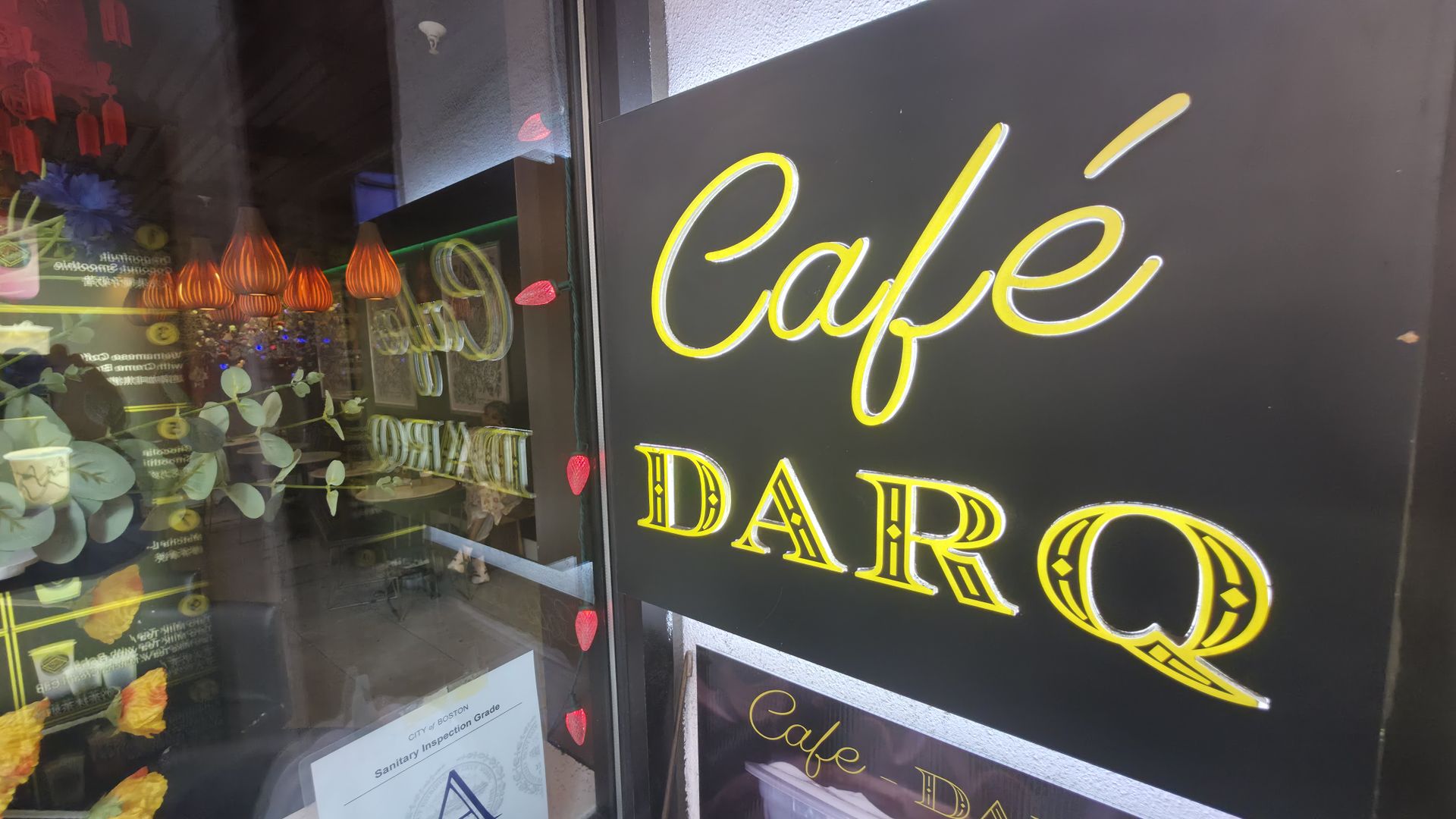 A storefront window with a large yellow neon-script "Cafe" on a black panel; through the glass, orange pendant lamps, green plants, and red heart decorations are visible inside.