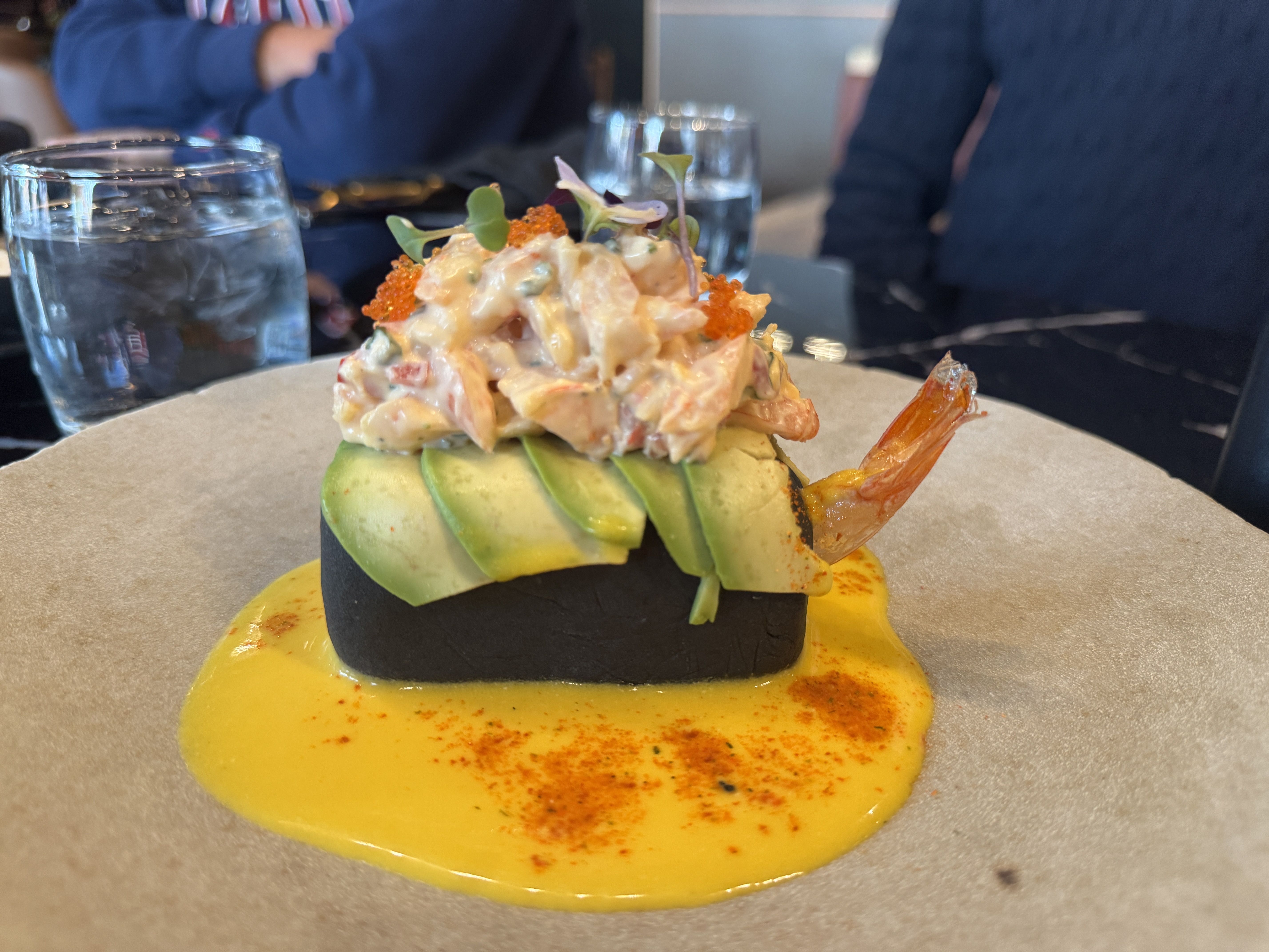 Gourmet dish with avocado slices, shrimp, creamy seafood salad topped with orange roe and microgreens, served on a black base with yellow sauce and sprinkled red spice on a beige plate.
