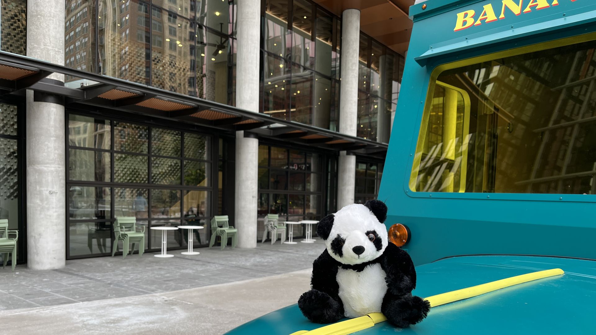 Go-Go, a stuffed animal panda bear, sits on the roof of a small green truck outside of a modern building with many windows.