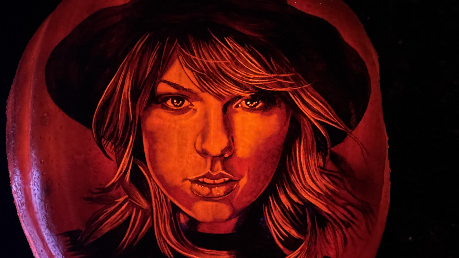 Close-up of an orange-painted female face on a red circular object, lit by red light. A hood surrounds the edge; the eyes and lips are visible with an intense gaze.
