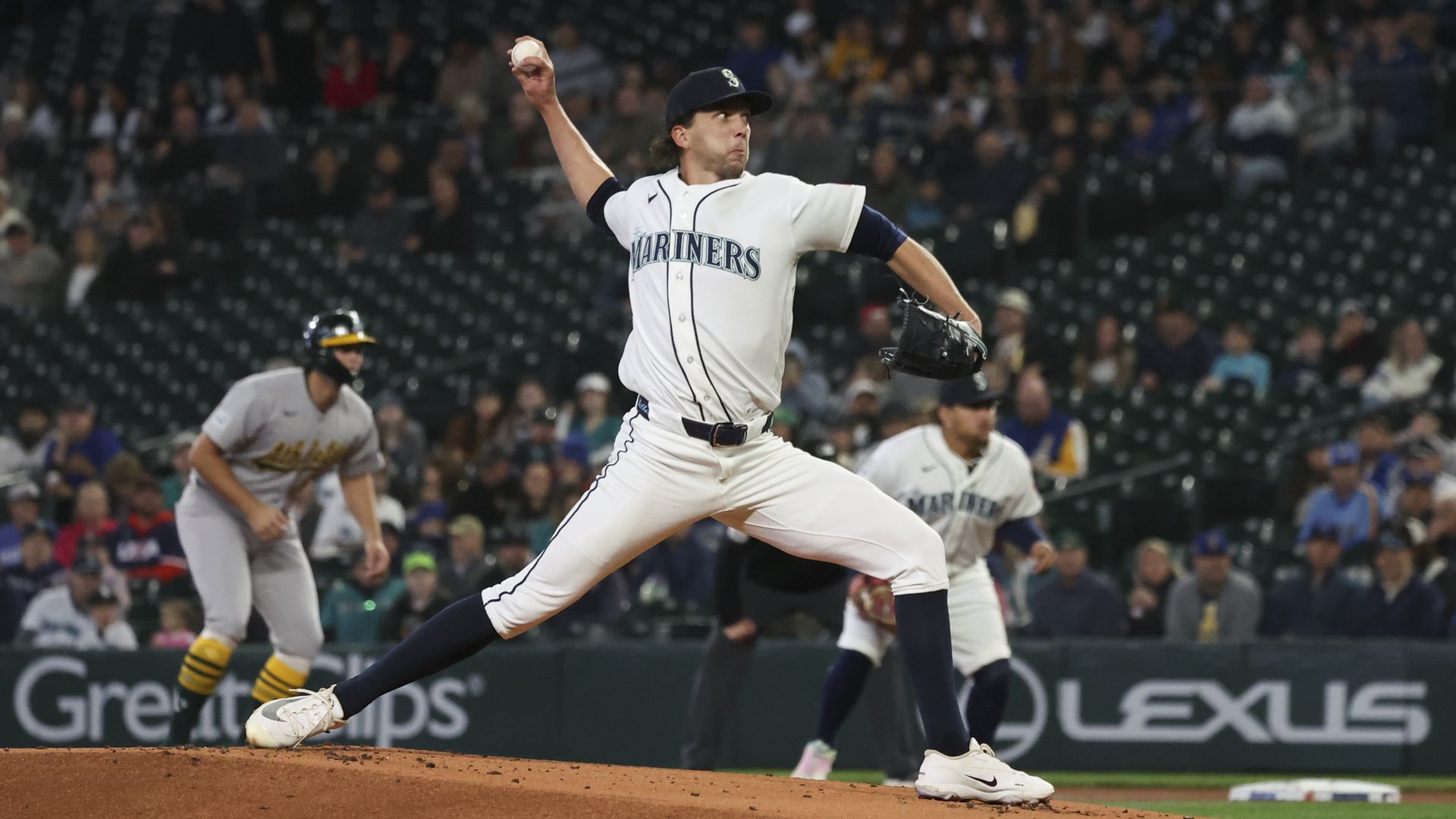 Mariners pitcher Logan Gilbert windes up for a pitch, with two players visible behind him and a crowd in the stands.