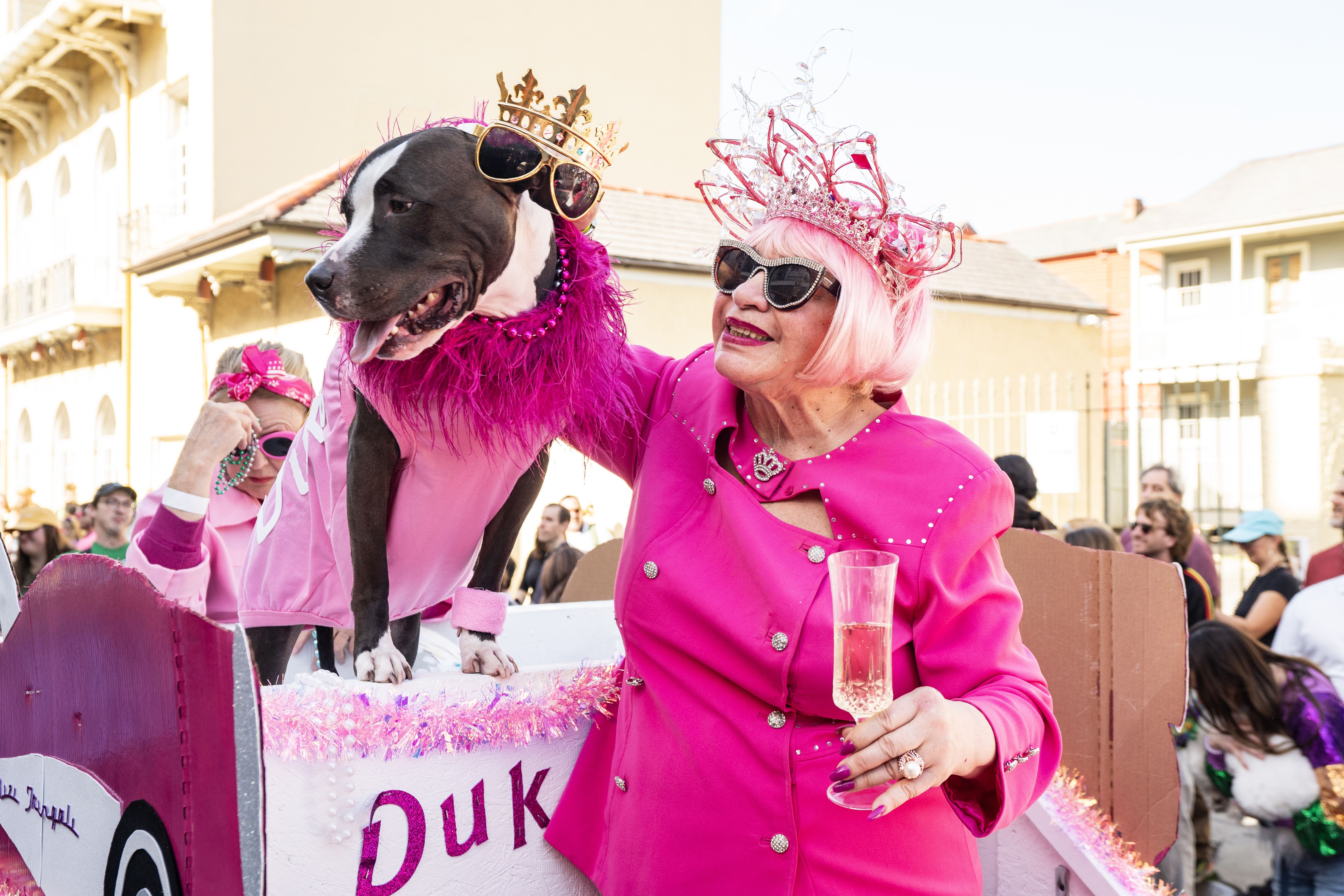 A woman holds a glass of champagne and pets her dog. Both wear crowns.