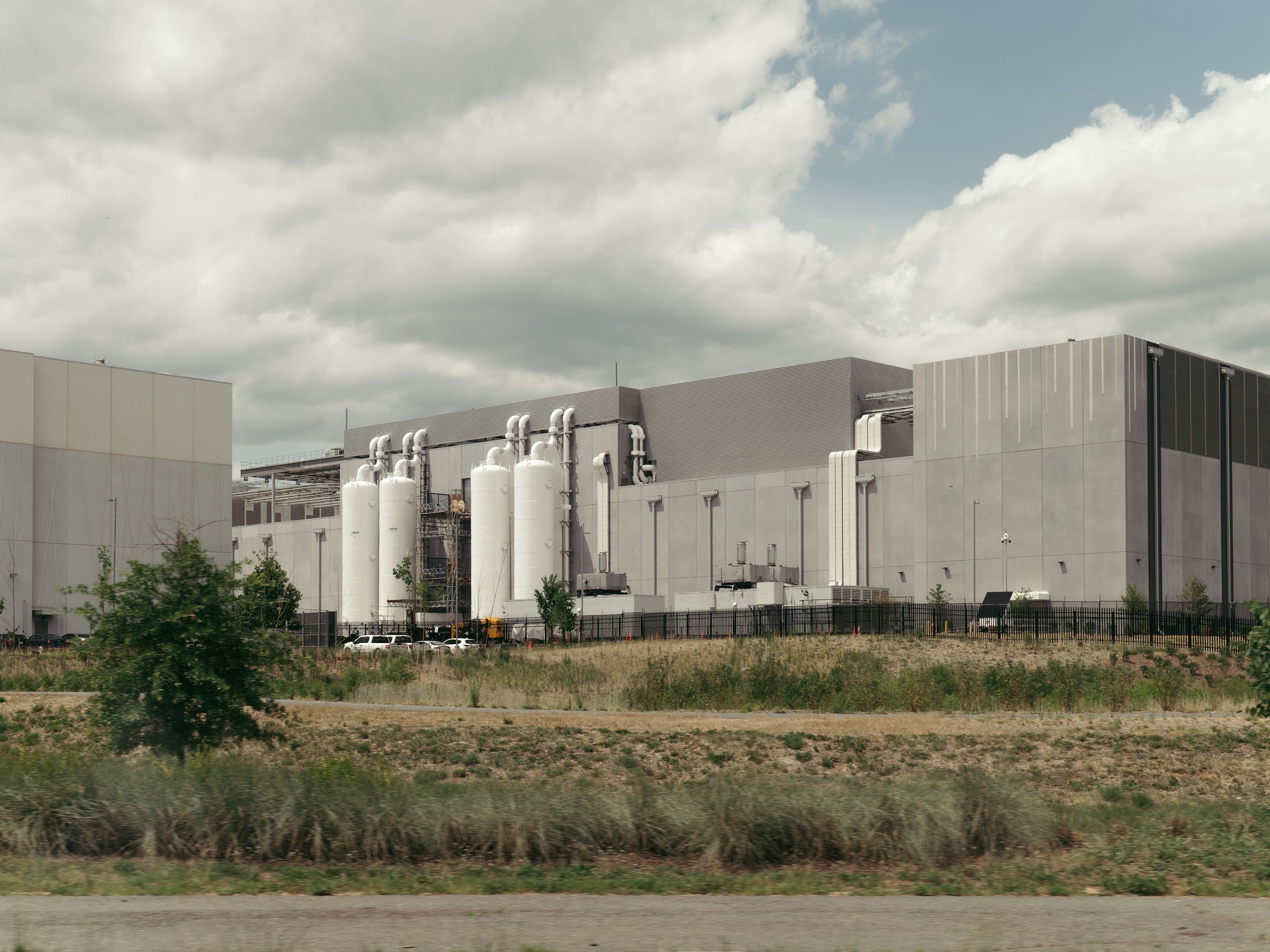 Industrial complex with gray buildings, white cylindrical tanks and metal pipes along the walls; a black fence in front, patchy grass and shrubs, and a cloudy blue sky overhead.