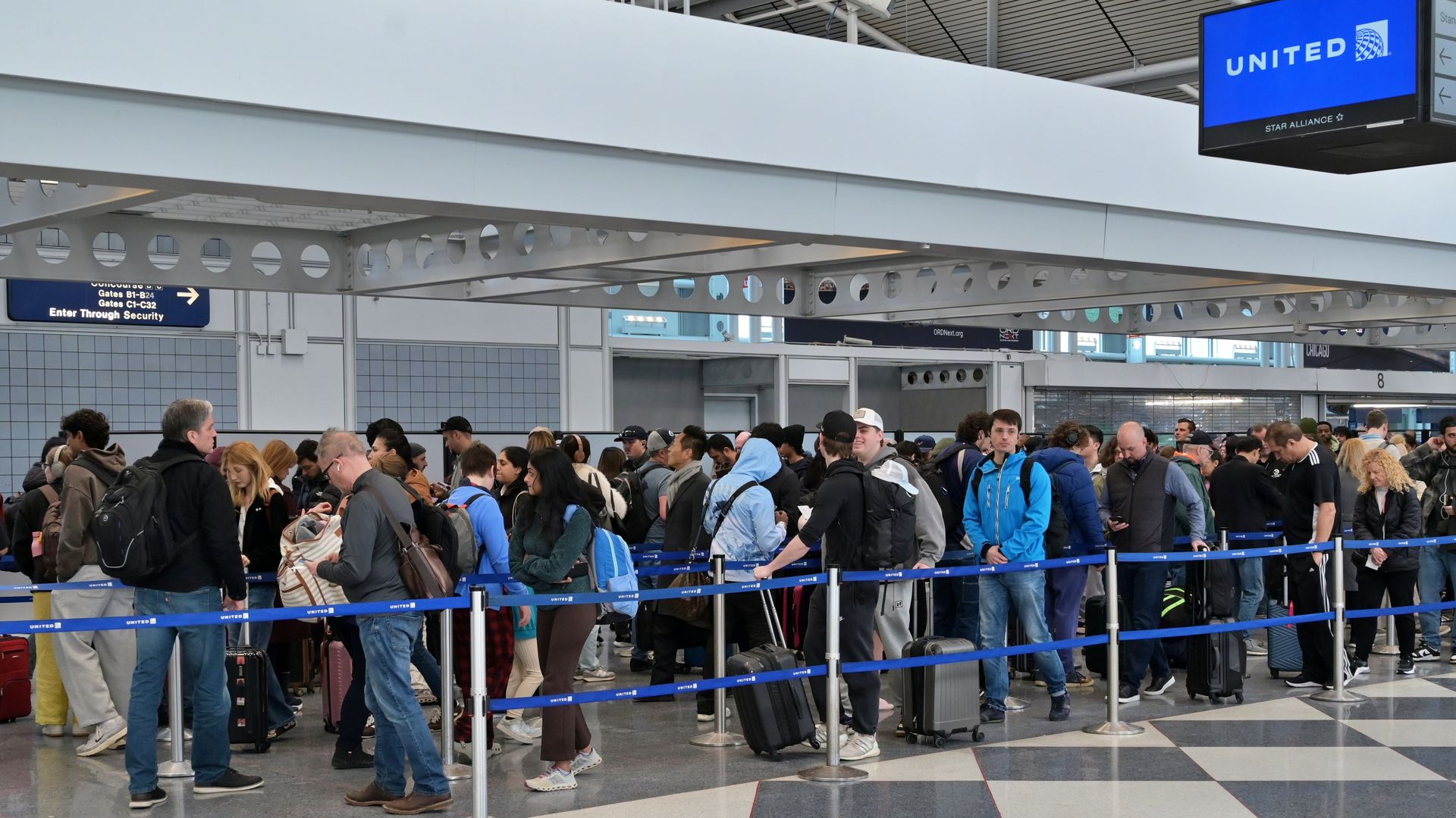 Long queue of travelers with luggage lined up behind blue crowd-control ropes at an airport check-in/security area, with a large overhead beam and a blue United sign overhead.