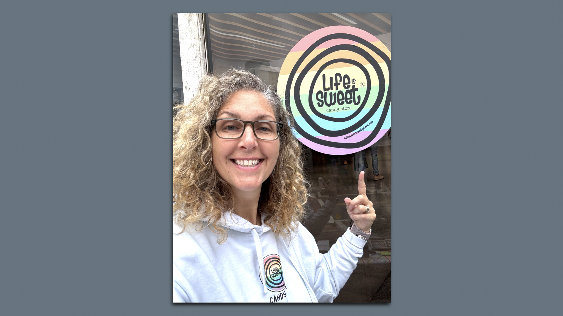 Smiling woman with curly hair and glasses points to a pastel rainbow candy store sign that says "Life is Sweet"; she wears a matching white hoodie with the same logo.