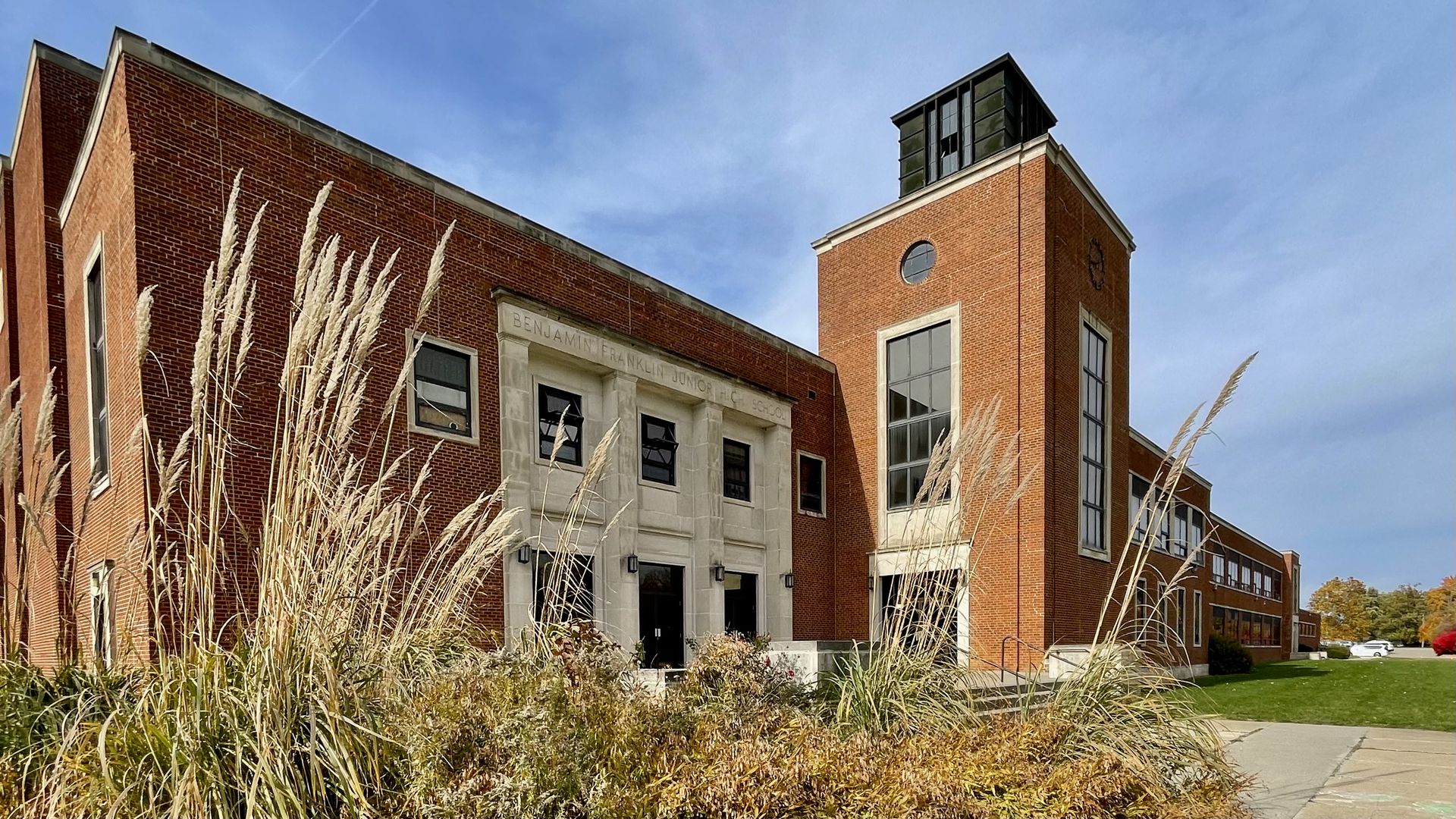 Brick building with tall windows and a clock tower labeled "Benjamin Franklin Junior High School," surrounded by tall grass and bushes under a clear blue sky.