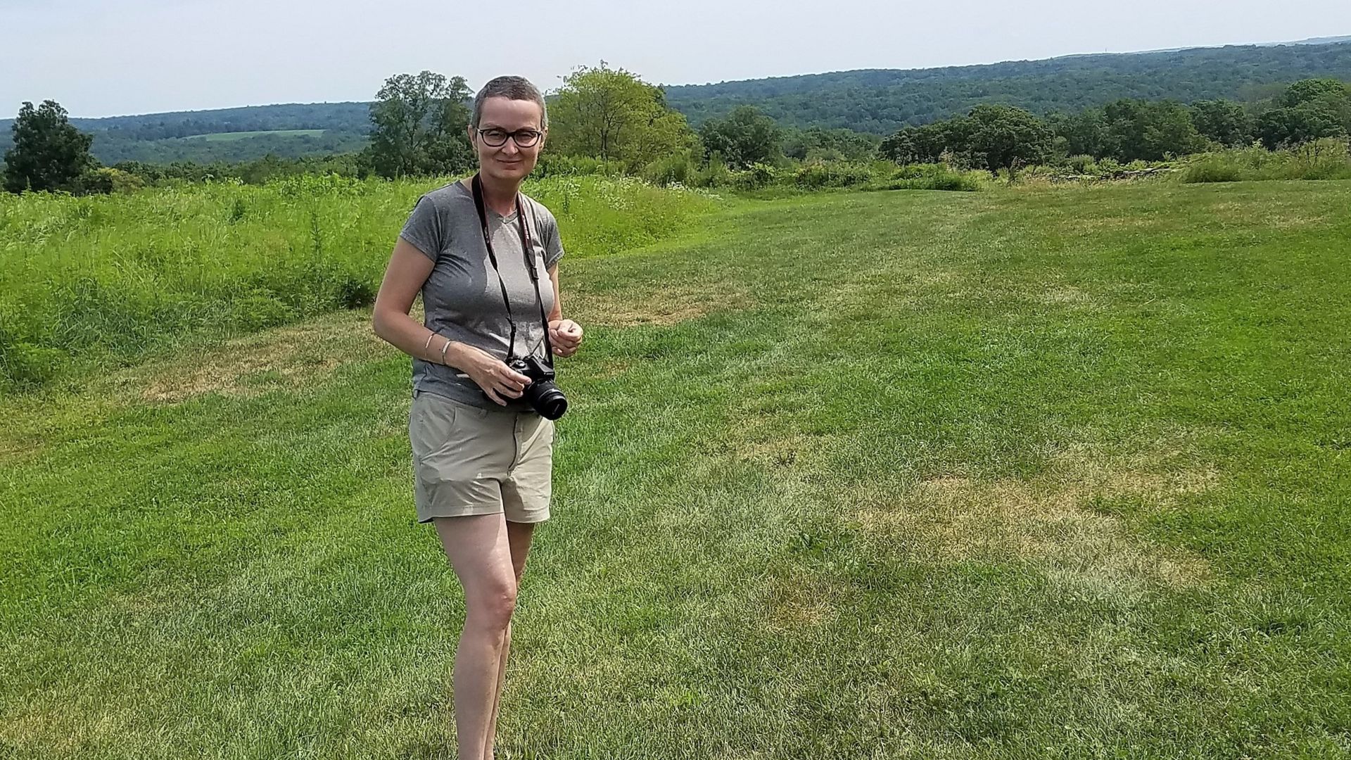 A woman stands in a green field on a sunny day, posing for a picture with a camera around her neck.