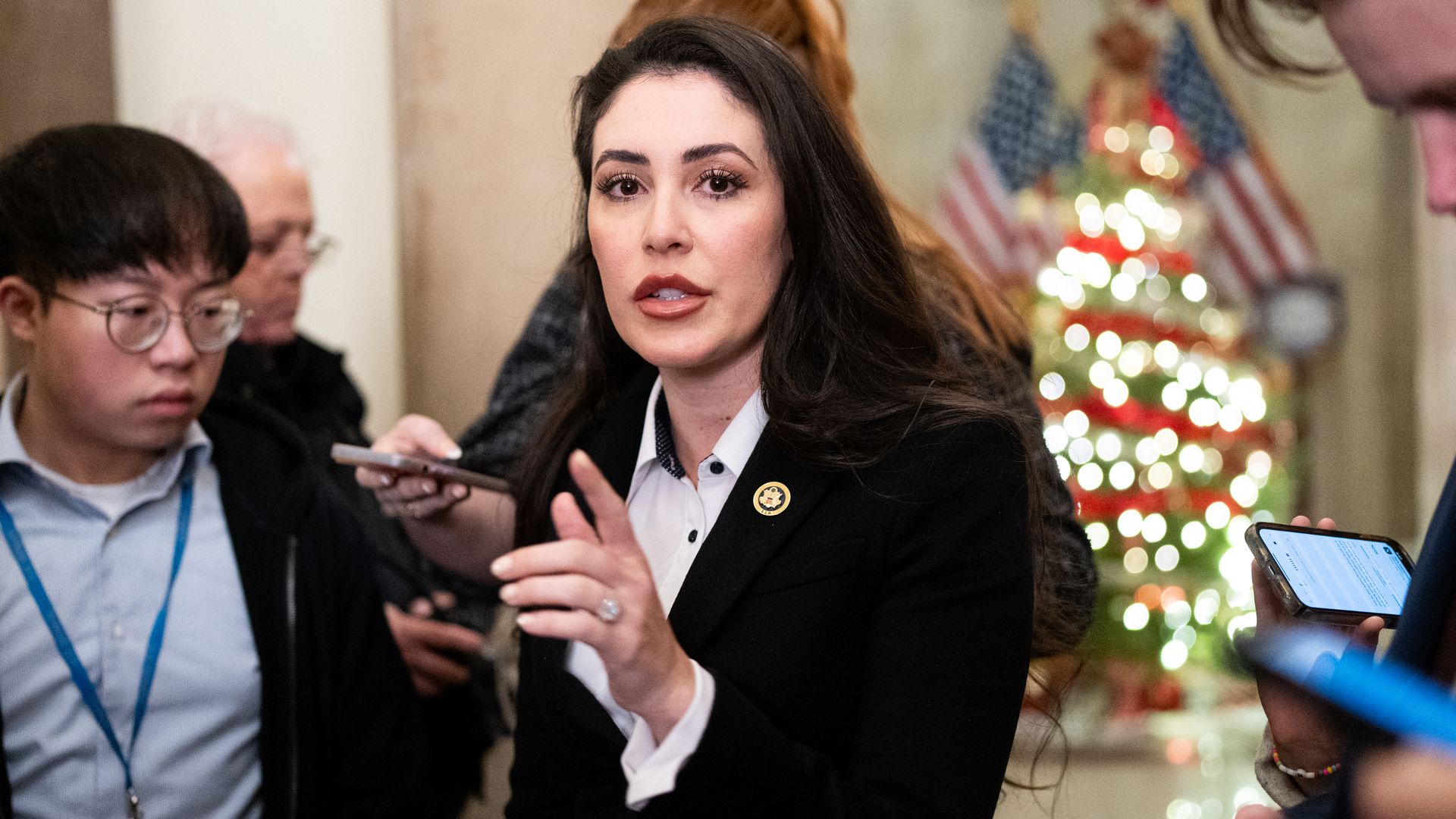 Anna Paulina Luna speaking to reporters while wearing a black blazer and standing in front of a christmas tree.
