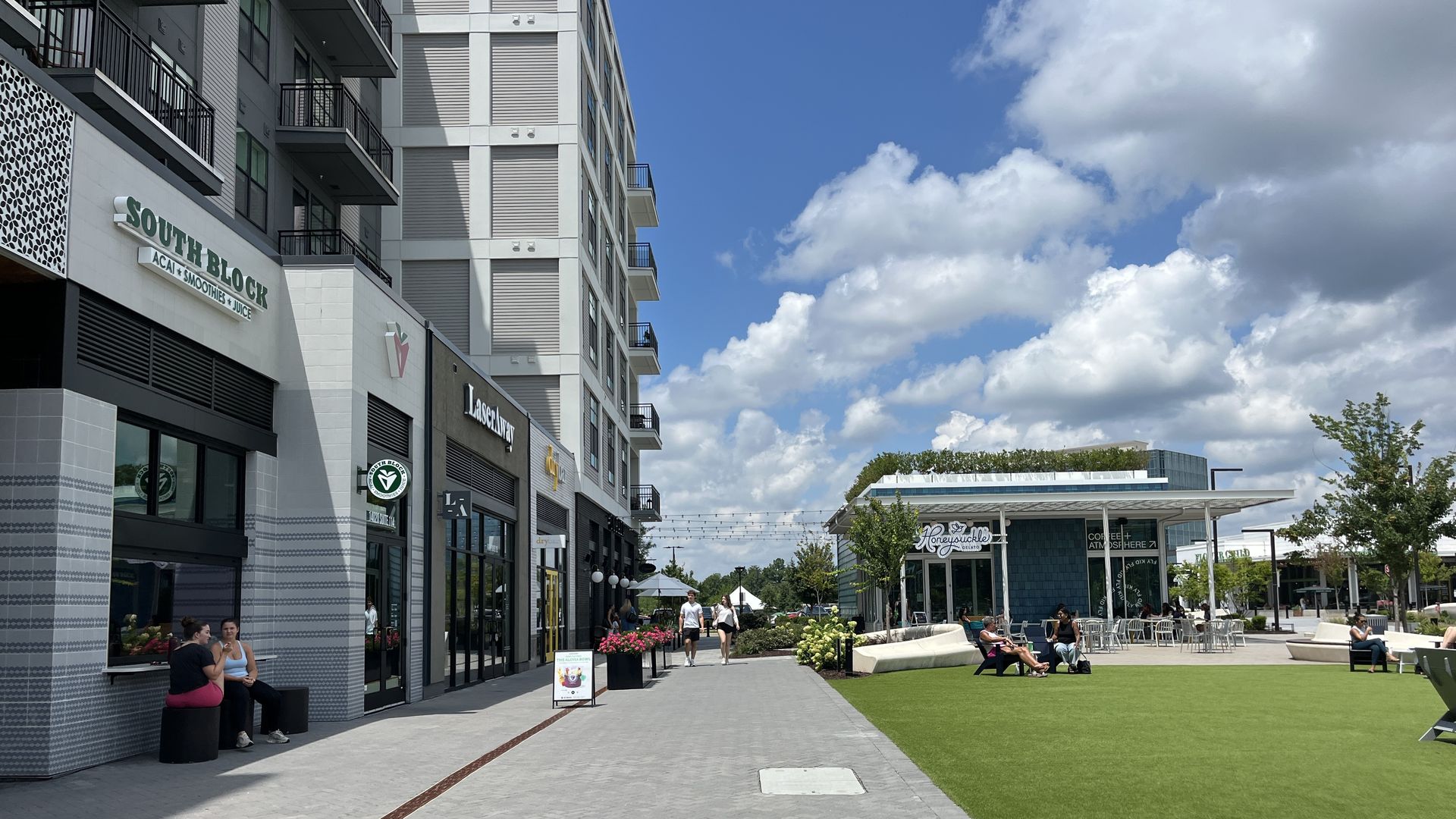 A modern, multi-story building with balconies on the left, a South Block storefront, and outdoor seating. A wide pedestrian plaza with a green lawn and people under a blue sky with fluffy clouds.