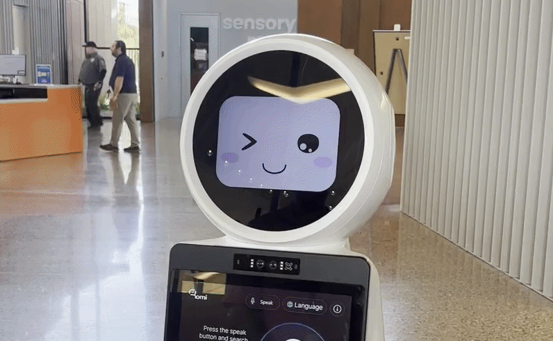 A moving video of a robot in the foreground winking and then smiling. People passing by in the background.