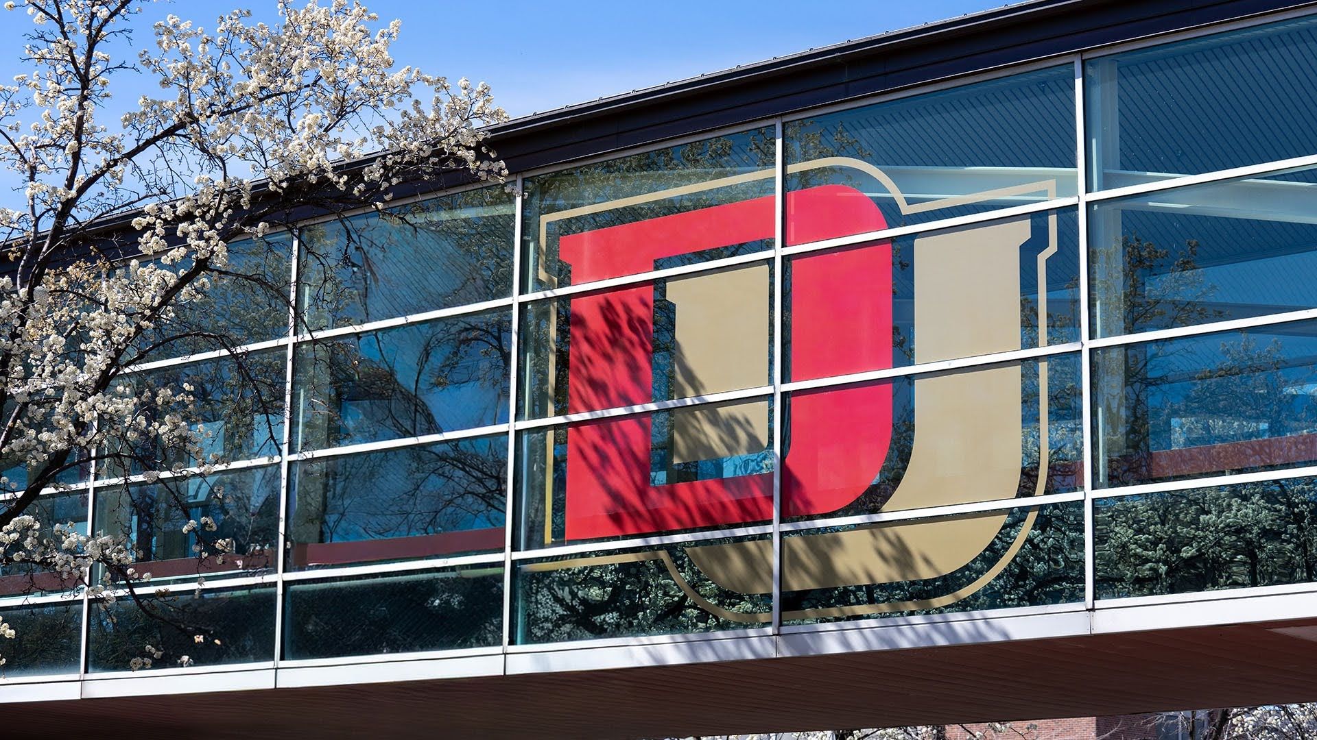 Glass building facade with a large red and beige geometric logo across the windows; blue sky reflections and white flowering tree branches in the foreground.