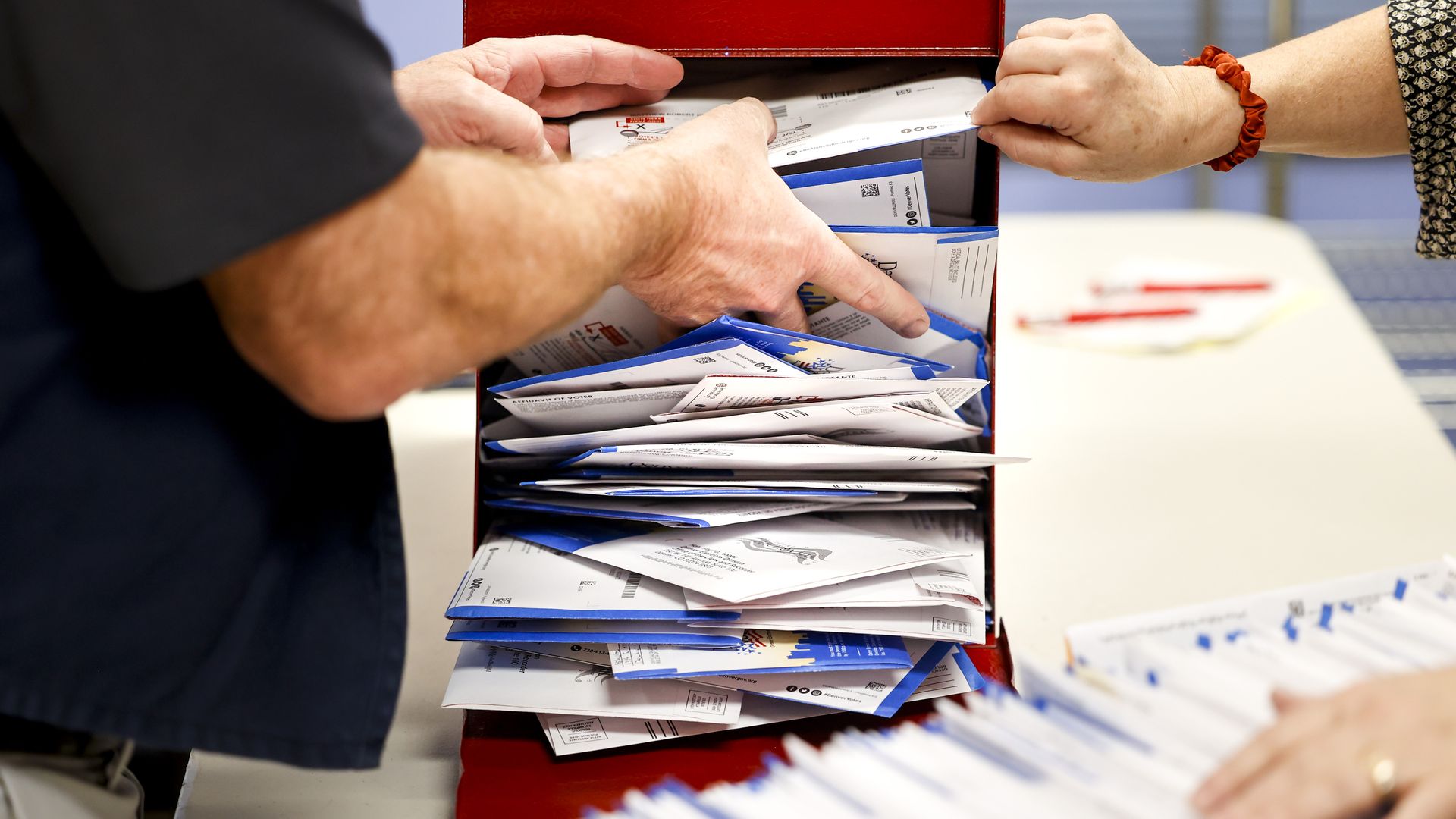 A pair of hands can be seen grabbing numerous ballots from a red metal container. 