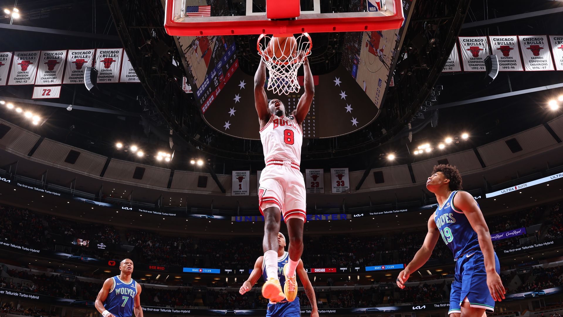 Chicago Bulls player in white jersey #8 dunks the ball through the hoop as blue-clad defenders watch, with Bulls banners and a crowded arena in the background.