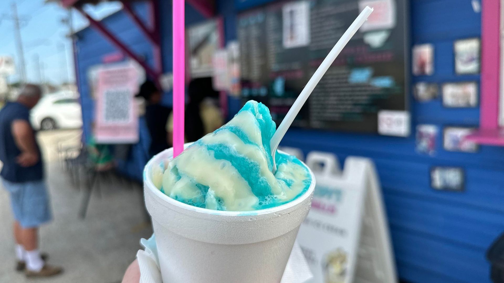 Photo shows a blue snoball in a white styrofoam cup with a pink straw and white plastic spoon. A blue and pink snoball stand is in the background.