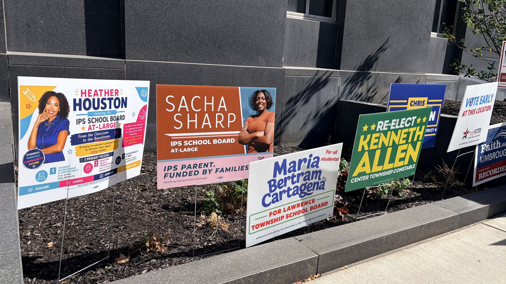 Row of colorful campaign signs along a sidewalk in front of a dark gray stone wall, featuring Heather Houston, Sacha Sharp, Kenneth Allen, Maria Bertram Cartagena, and other candidates.