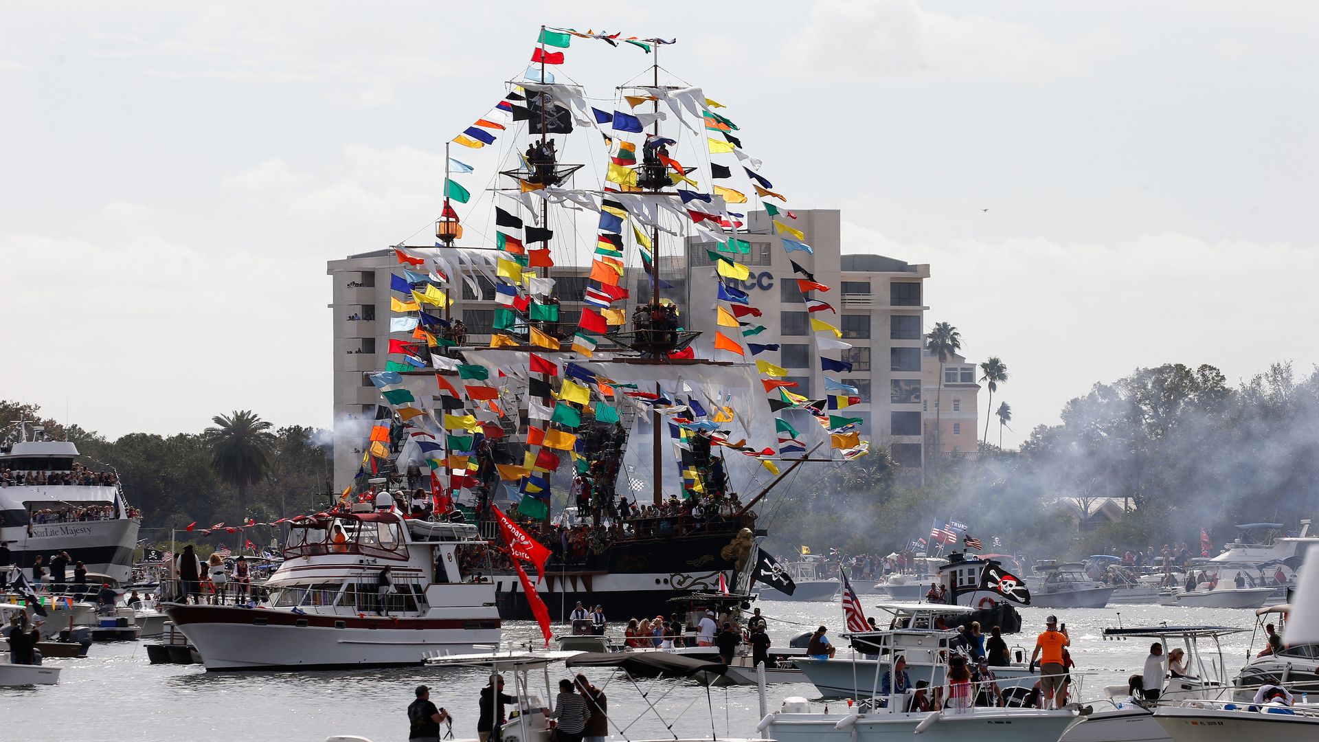 A pirate ship with dozens of multi-colored flags along the masts surrounded by modern boats.