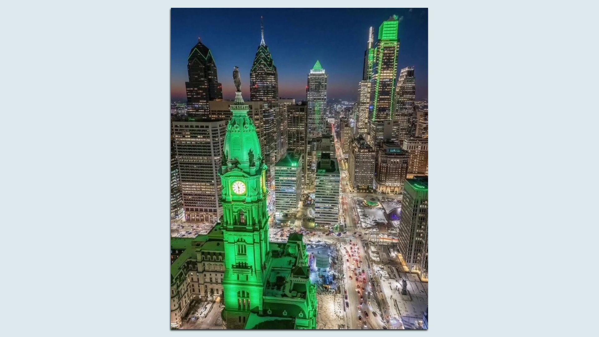 Nighttime Philly skyline with prominent clock tower lit in green, surrounded by skyscrapers also illuminated with green accents, busy streets filled with cars, and a snowy ground scene.