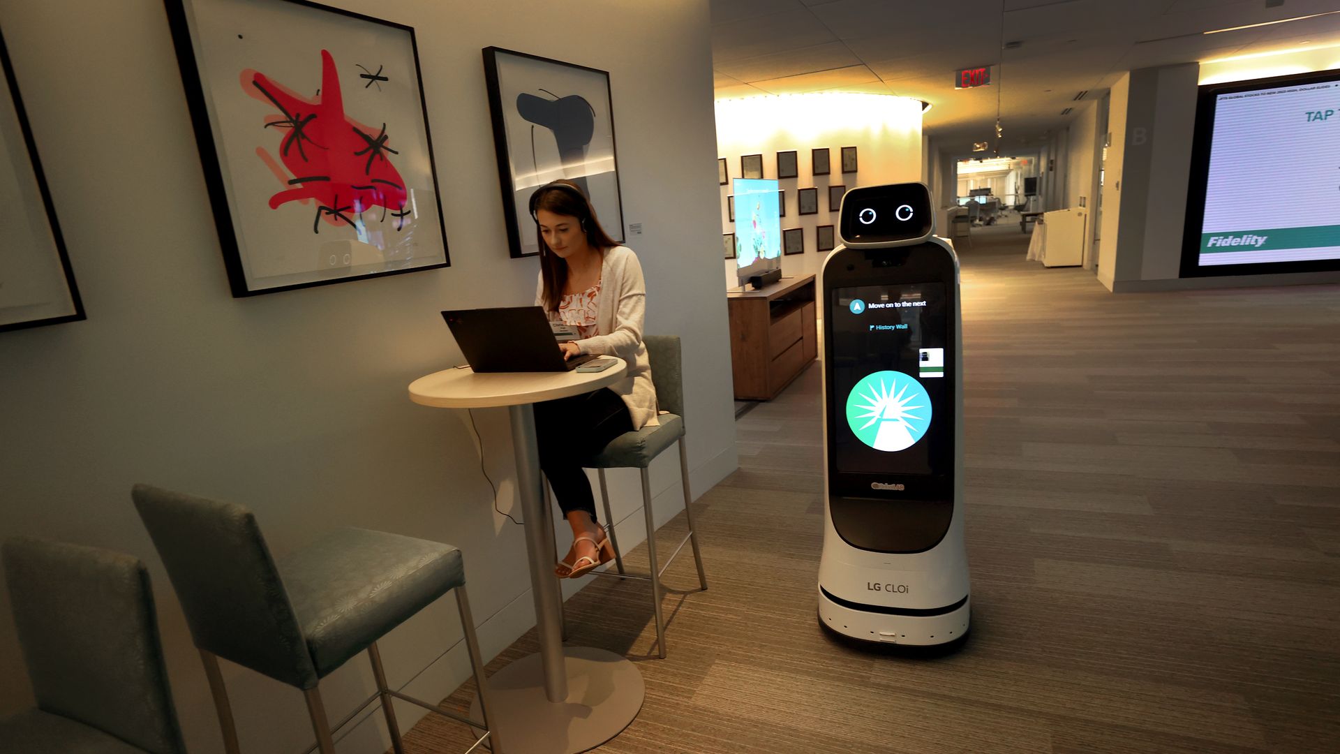 A meandering robot strolls by a woman seated in an office corridor.