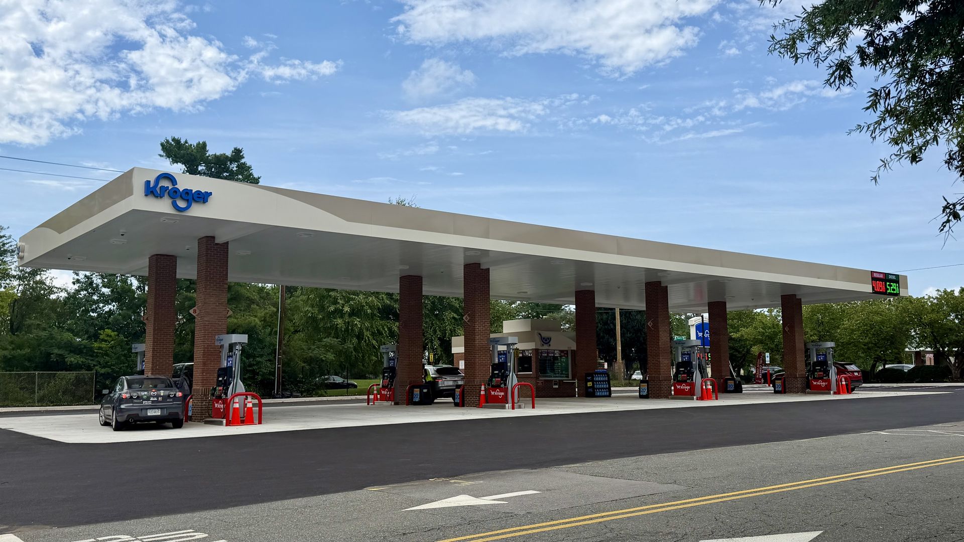 Wide shot of a Kroger gas station with a cream and tan canopy supported by brick columns, blue Kroger sign, multiple gas pumps, cars, and a partly cloudy blue sky.