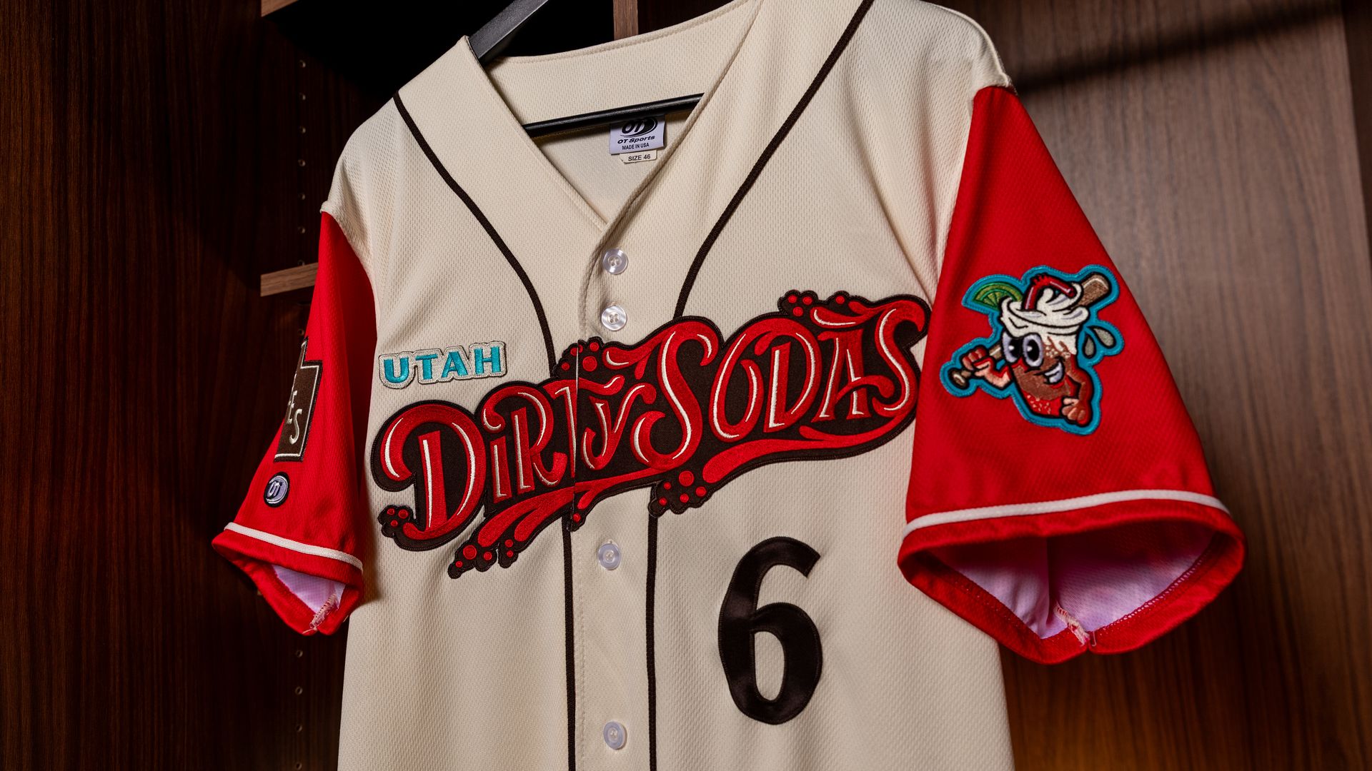 Cream and red Utah Dirty Sodas baseball jersey with number 6, hanging on a black hanger inside a wooden locker.