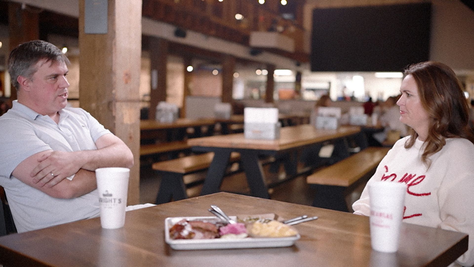 Two people sit at a wooden table in a casual restaurant. A man in a light blue shirt faces a woman in a white sweater; a plate of food and cups sit on the table, with a blurred background.
