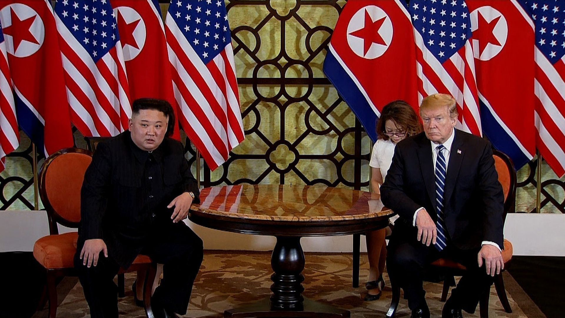 In this image, Donald Trump and Kim Jong Un sit across from each other at a round wooden table, with the American and North Korean flags draped behind them.