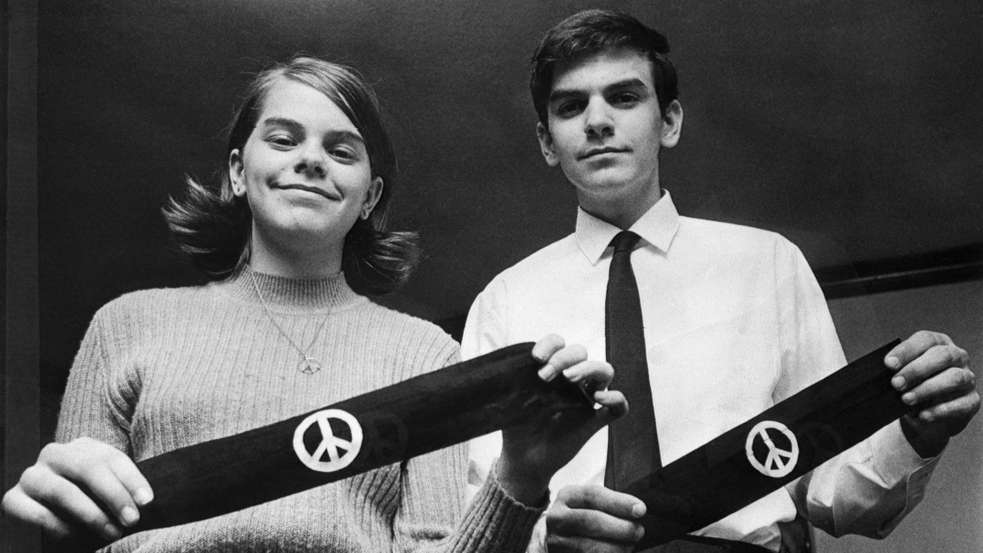 Black and white photo of a smiling young woman in a sweater and a young man in a white shirt and tie holding black armbands with white peace symbols.