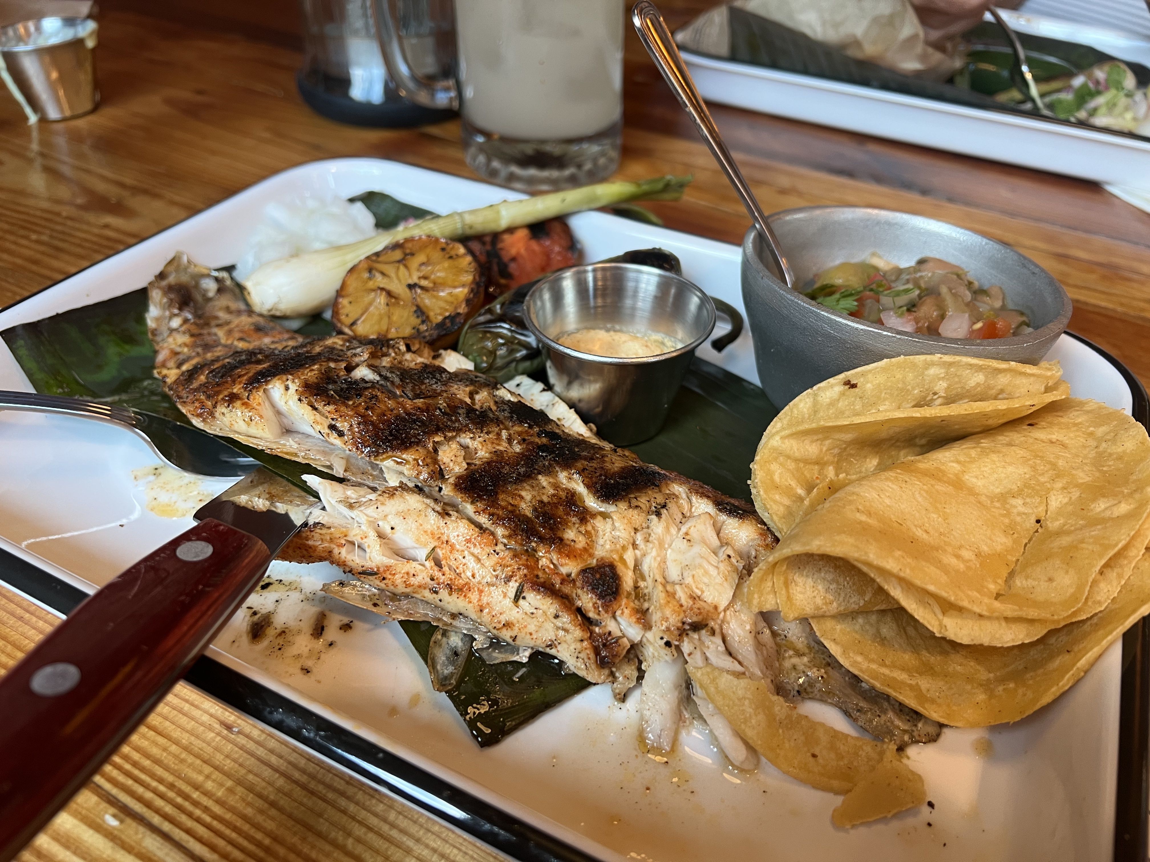 Grilled fish with charred lemon, grilled vegetables, salsa, crema in a metal cup, and a stack of folded yellow corn tortillas on a white tray with a knife and spoon on a wooden table.