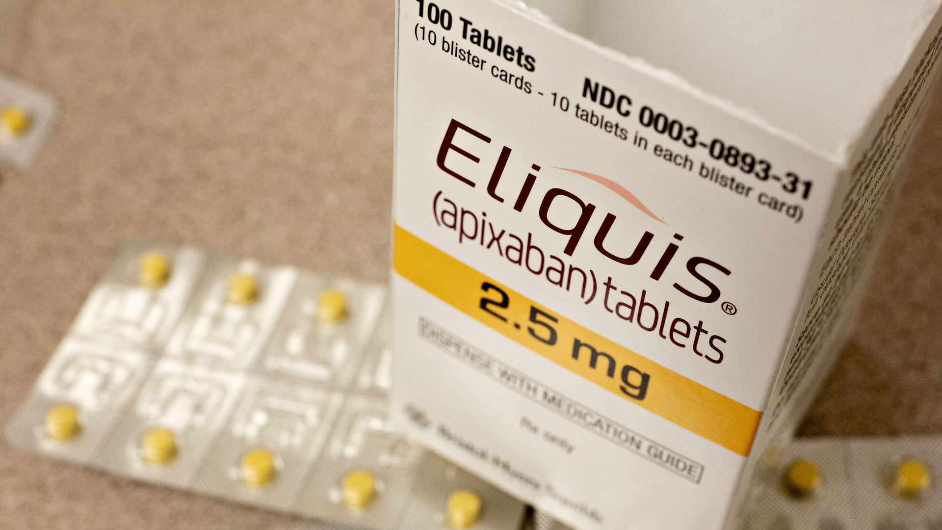 A white Eliquis box with yellow Eliquis tablets spread out underneath the box.