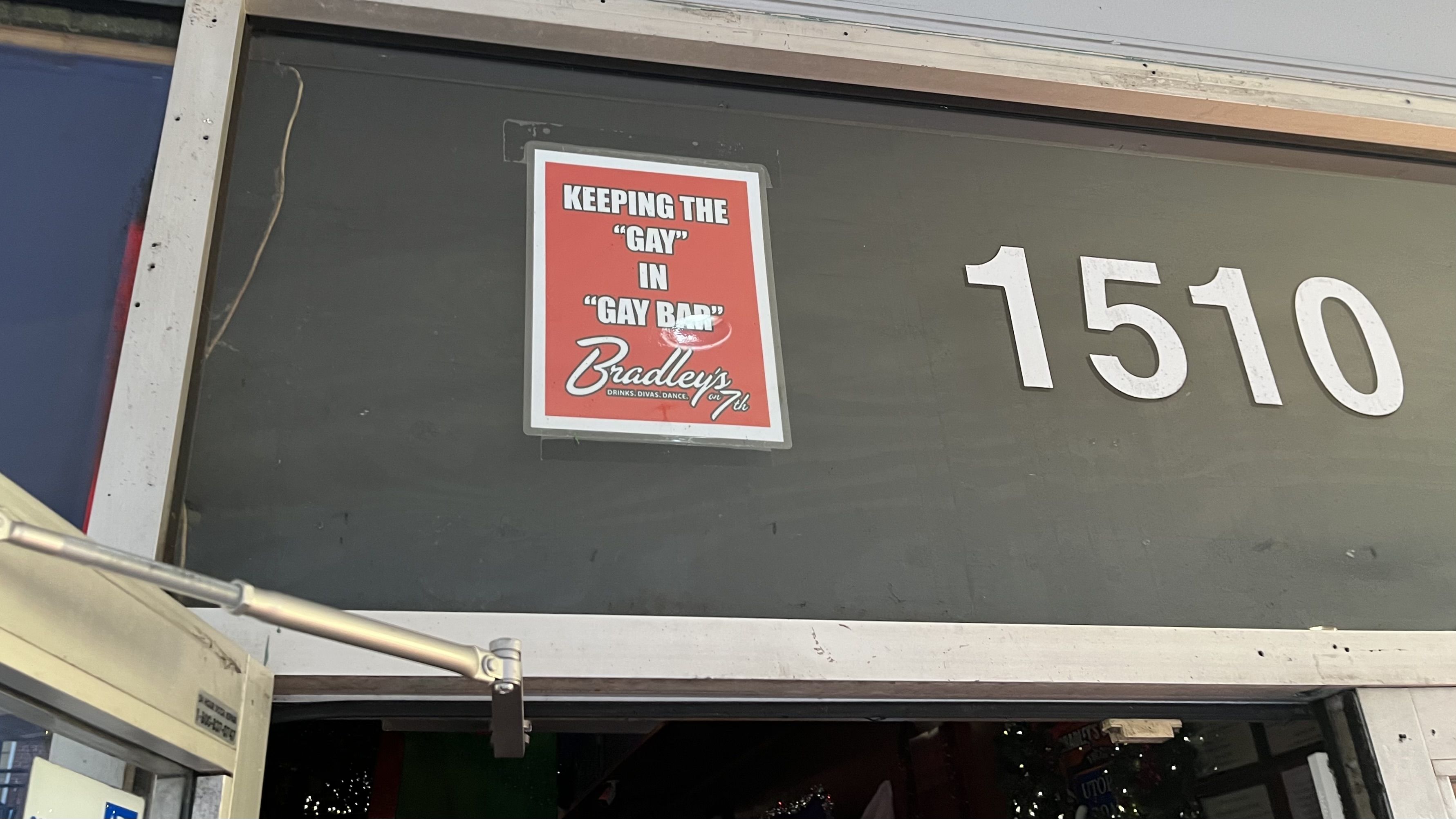 Sign with red background on a dark door, saying KEEPING THE "GAY" IN "GAY BAR" above Bradley's on 7th logo, next to white numbers 1510.